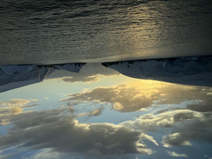A landscape image features a body of water with sunlight reflecting on its surface. Above, a sky with clouds in various shades of gray and white. The horizon line appears jagged and irregular, with dark, snow-covered landmasses. The overall perspective is unusual, with the water seemingly above the sky and land, creating a surreal, flipped orientation. The colors are muted, consisting mostly of blues, grays, and whites with golden light highlighting the water and clouds.