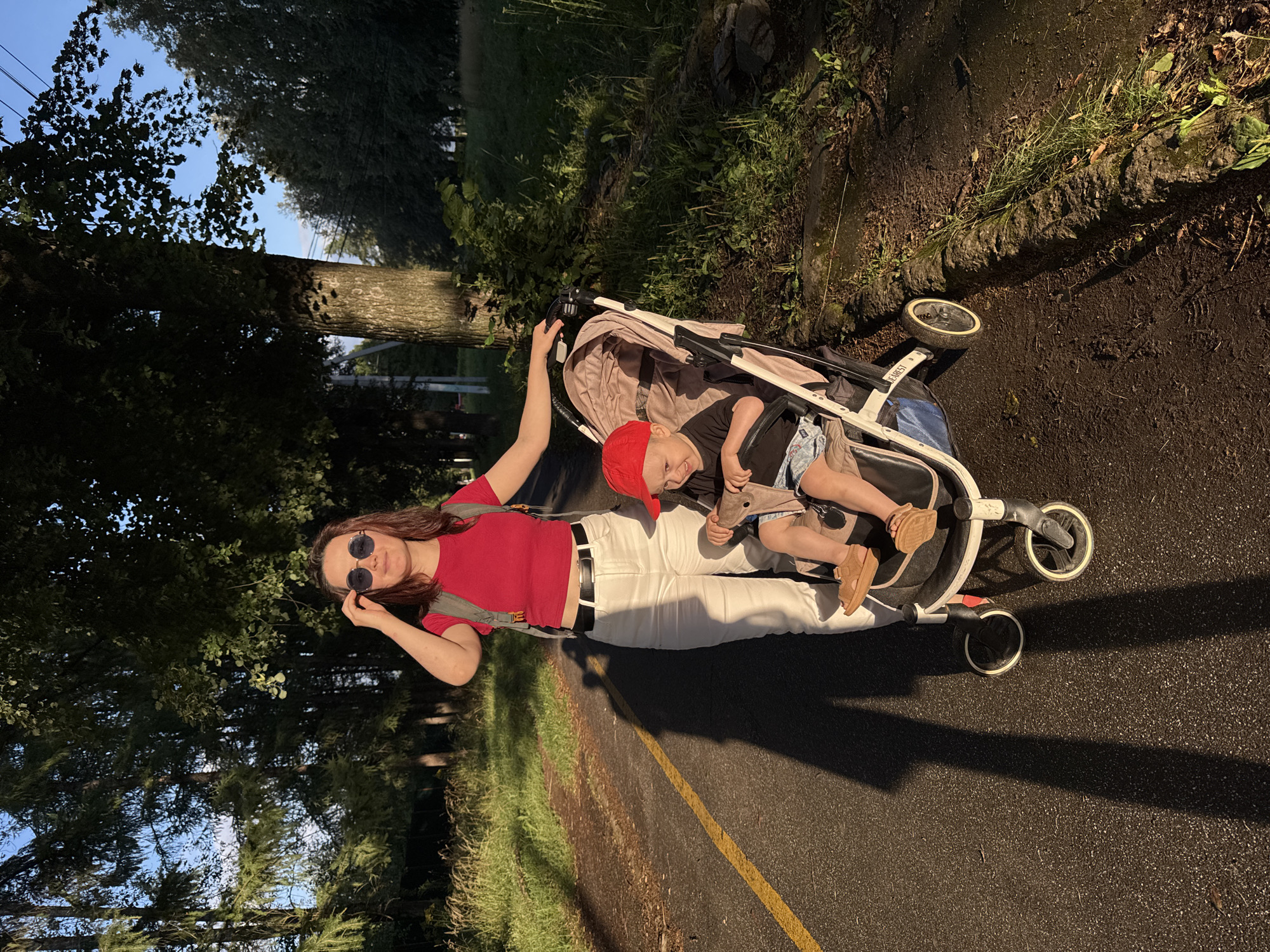 A woman with shoulder-length brown hair, wearing sunglasses, a red crop top, and white pants, stands beside a stroller on an asphalt path. She is holding onto the stroller's handle. Inside the stroller, a young child wearing a red hat, black t-shirt, and patterned shorts sits, looking to their right. The path is bordered by grass and trees on the right and grass and trees on the left. A yellow line marks the edge of the asphalt path. The scene is illuminated by the golden light of what appears to be sunset. This image was taken in Izhevsk, Russia.