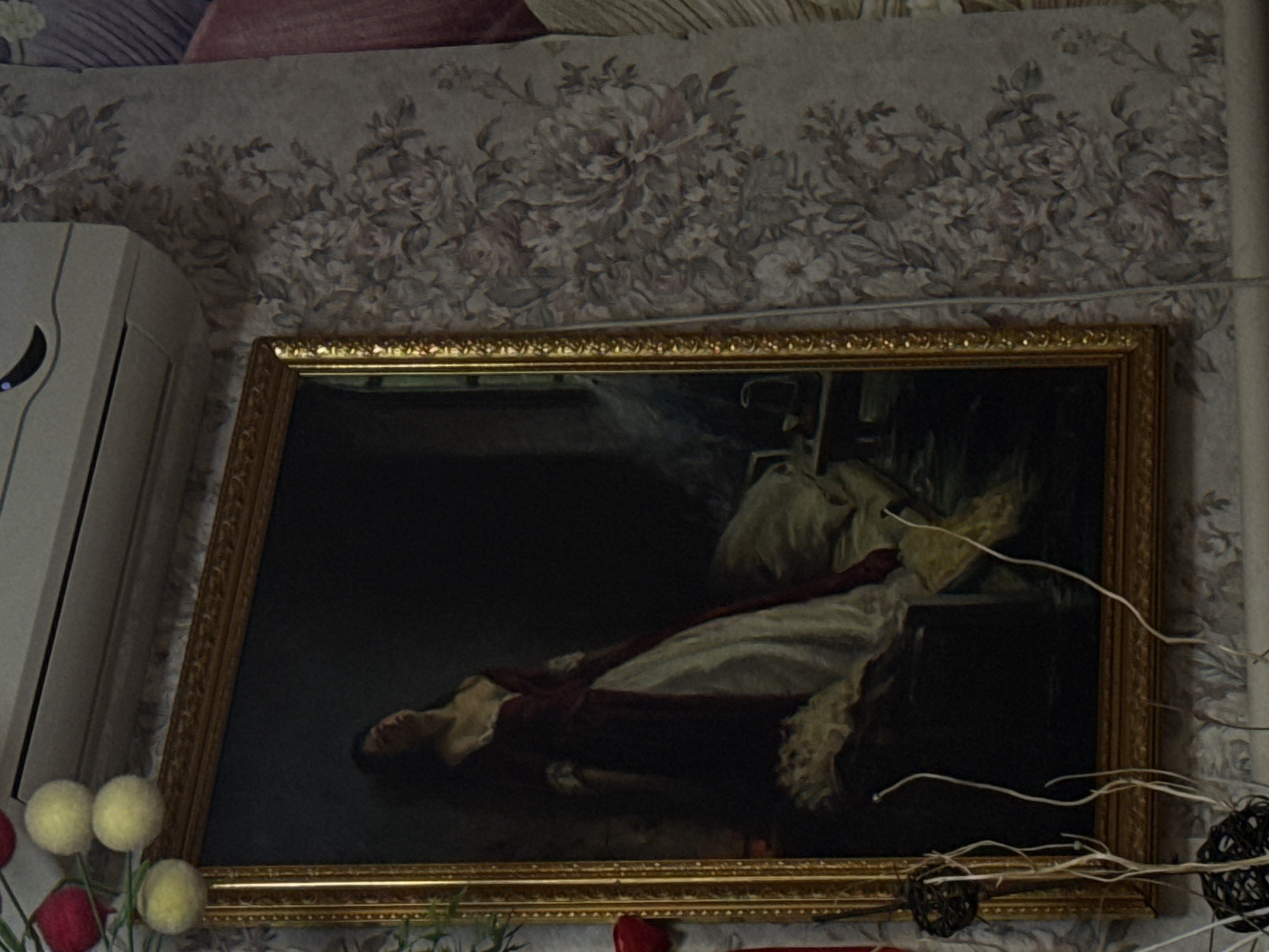 Here's a description of the image:
The image shows a framed painting hanging on a wall with floral wallpaper. The painting features a woman in a dark, romantic style. She is lying down, wearing a white dress with lace details and a deep red overlay. The backdrop is dark, with some lighter areas that may represent drapery or other furnishings. A gold frame surrounds the artwork. To the left of the painting is an air conditioner unit. In the bottom left corner, there is a decorative floral arrangement with what appear to be fuzzy yellow and red elements. A dried decorative branch is located on the right.