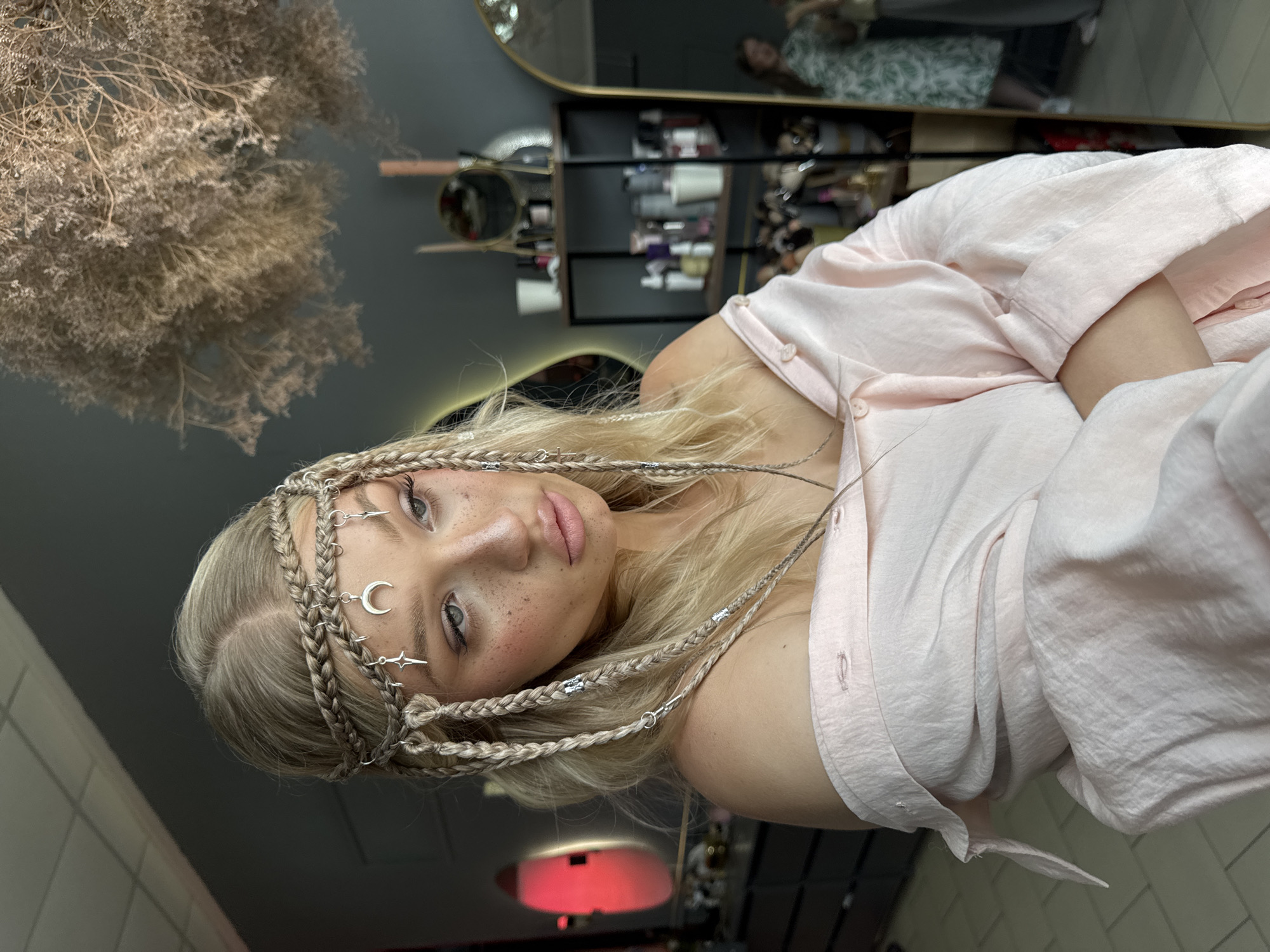 A young woman with blonde hair styled in braids and adorned with celestial-themed hair jewelry is shown in a close-up portrait. She is wearing an off-the-shoulder pale pink blouse and has makeup applied, including freckles. The background suggests an indoor setting, possibly a beauty salon or dressing room, with shelves containing various bottles and containers, mirrors, and decor elements like dried plants. There is no information available to confirm the location as Novocherkassk, Russia.