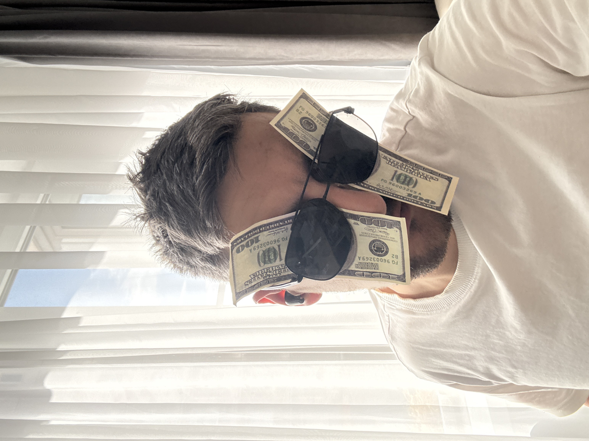 A man with grey hair is wearing sunglasses with three one hundred dollar bills stuck to them, blocking his eyes. He is wearing a white t-shirt. In the background, there are window blinds, with some daylight filtering through.