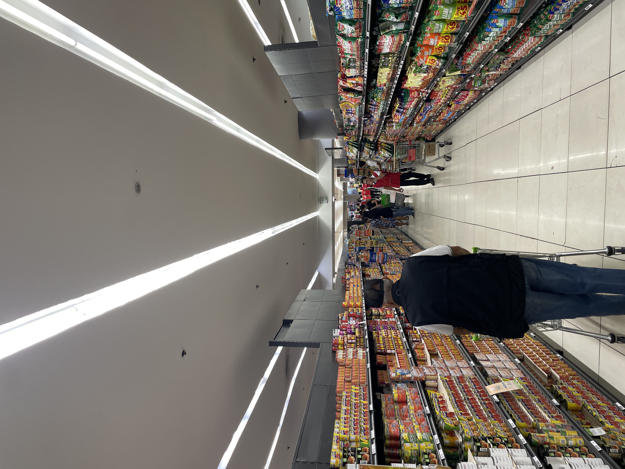 The image depicts a wide, well-lit aisle in a supermarket, likely located in Davao City, Philippines, judging by the presence of products and signage that are common in the region. The aisle is lined with tall shelves stocked with a variety of canned goods and packaged snacks on both sides. The floor is tiled in a checkered pattern of white and light grey.
In the foreground, a man wearing a black vest over a white shirt and blue jeans pushes a shopping cart. He is walking away from the camera, deeper into the supermarket. His face is not visible, but he appears to be an elderly gentleman.
Further down the aisle, several other shoppers and supermarket employees can be seen. To the left, a group of people are browsing the shelves. To the right, a supermarket employee in a red uniform is visible, potentially assisting customers. The ceiling features long, bright fluorescent light fixtures, illuminating the products and the general space. The overall atmosphere is that of a typical busy retail environment.
