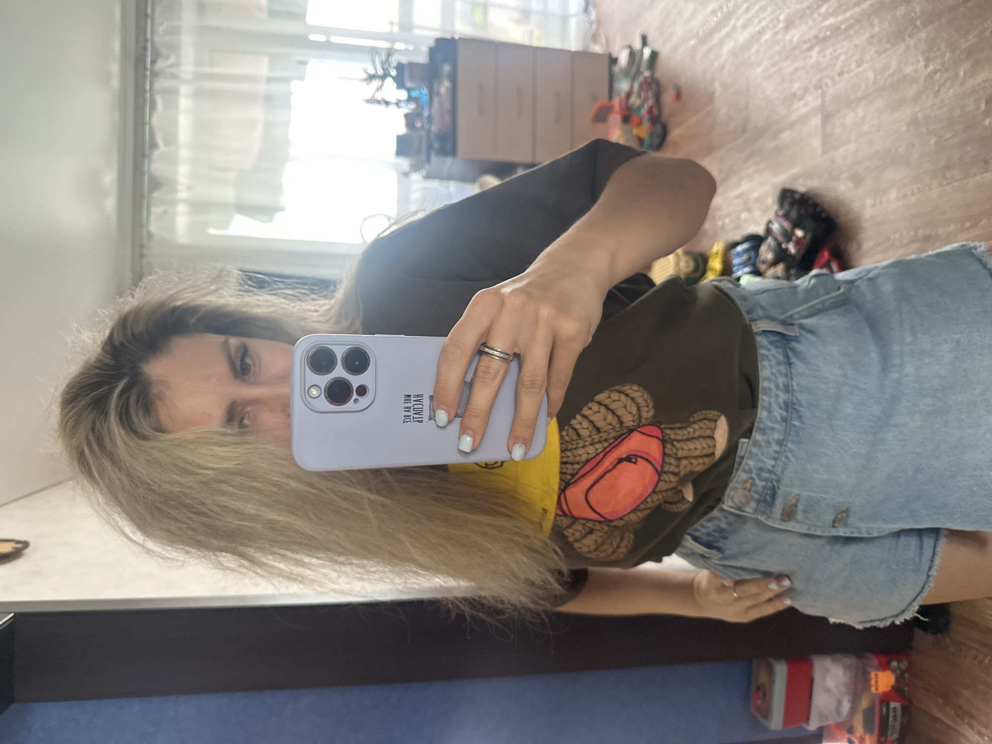 A woman, reflected in a mirror, holds a light purple phone to take a selfie. She has long, blonde hair that partially obscures her face. She is wearing a dark t-shirt with a graphic of a teddy bear and light blue denim shorts. The t-shirt appears to have text on the sleeve. Her fingernails are painted a pale blue. She is wearing a ring on her left hand.
The background, also seen in the mirror's reflection, shows a room with wooden flooring and a dresser with drawers. Various toys are scattered on the floor, and there is a window with blinds allowing sunlight to stream in. The setting appears to be a home environment, likely daytime due to the natural light. The overall mood is casual and relaxed. Some text is visible on the phone case and possibly on the sleeve of the t-shirt, but it is not legible. The image was taken in Novoaltaysk, Russia.
