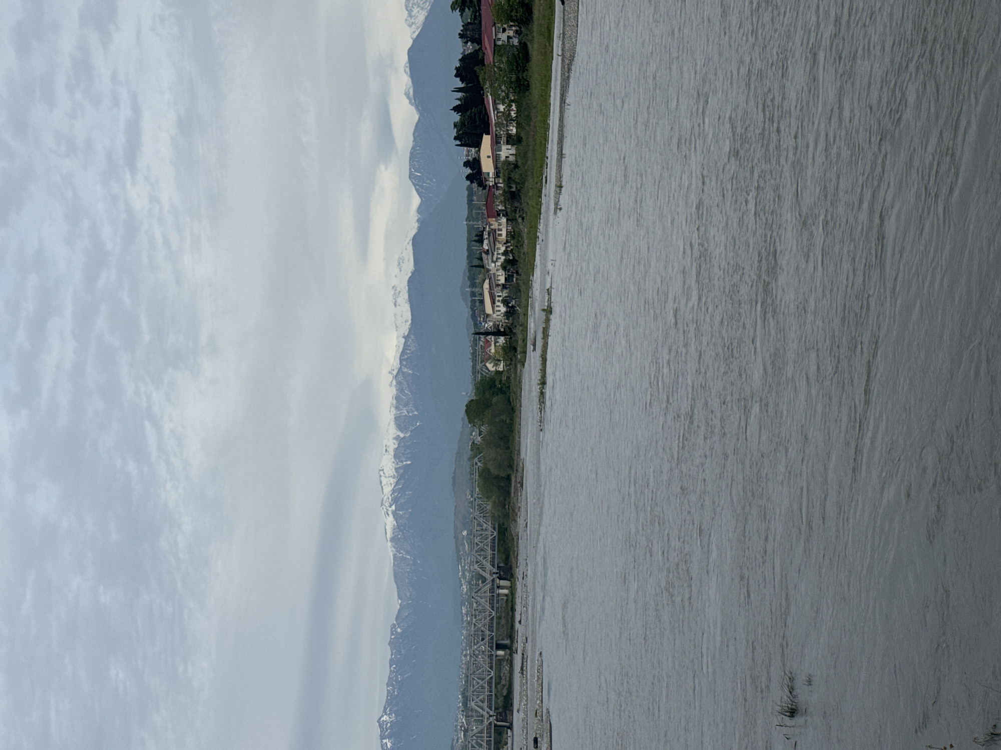 A landscape view shows a wide, gray river on the right, occupying about half the image. The river flows toward the bottom of the frame. On the left, tall mountains with snow-capped peaks stretch into the distance under a cloudy sky.
Along the riverbank, a narrow strip of land shows houses and buildings nestled among trees. A steel railway bridge crosses the river, connecting the riverbank to the mountainous terrain.
The photograph depicts an area near Sochi, Russia, characterized by its river valleys and mountain landscapes.