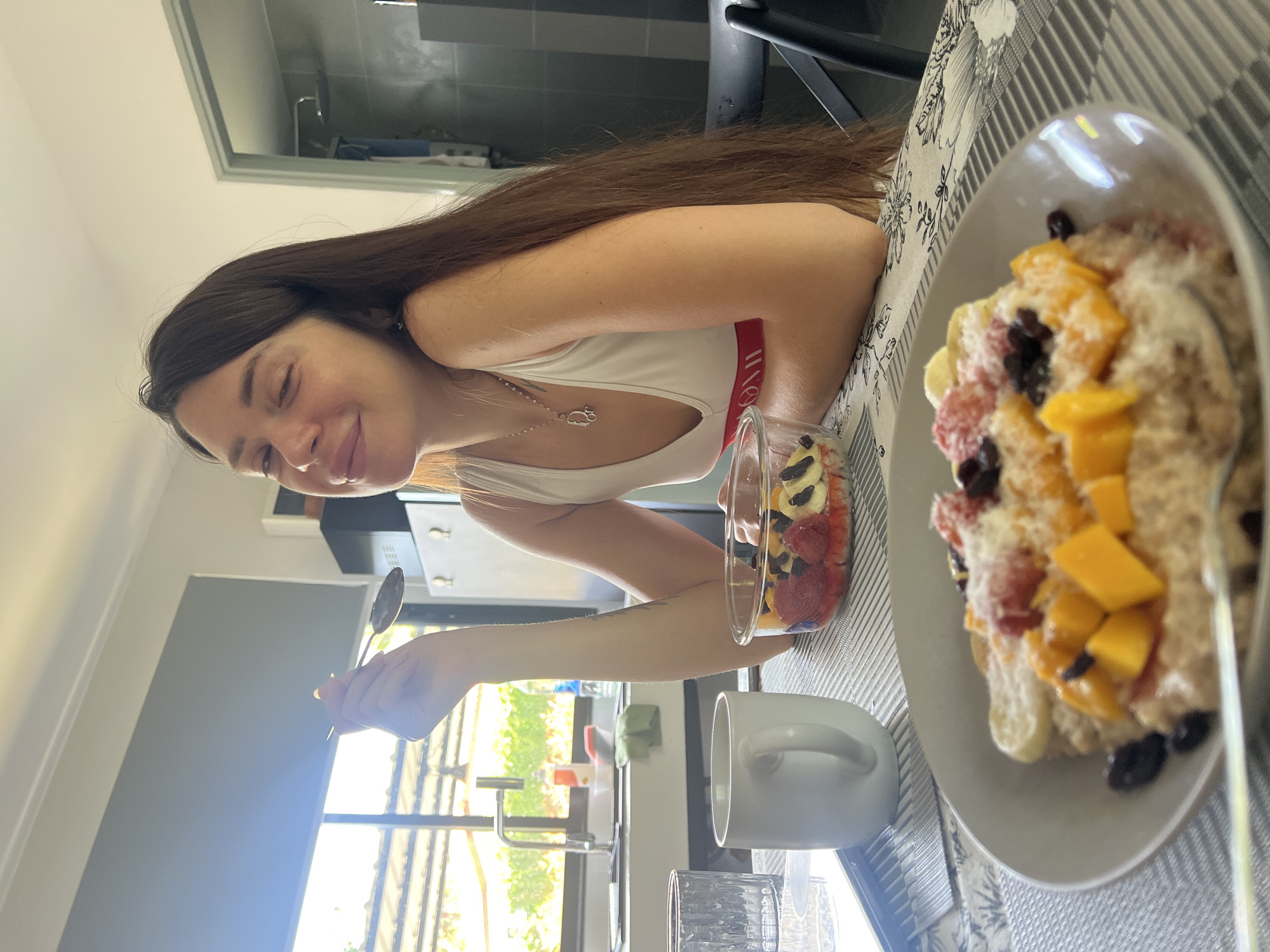 A woman with long brown hair is seated at a table, holding a spoon in her right hand, presumably about to eat. On the table are two bowls of fruit, oatmeal and various sliced fruits. A white mug and a transparent glass are also on the table. The setting appears to be indoors, possibly a kitchen, with a window visible in the background, revealing greenery outside. Given the prompt's specification, the location is in the city of บ้านบางรักษ์, Thailand.