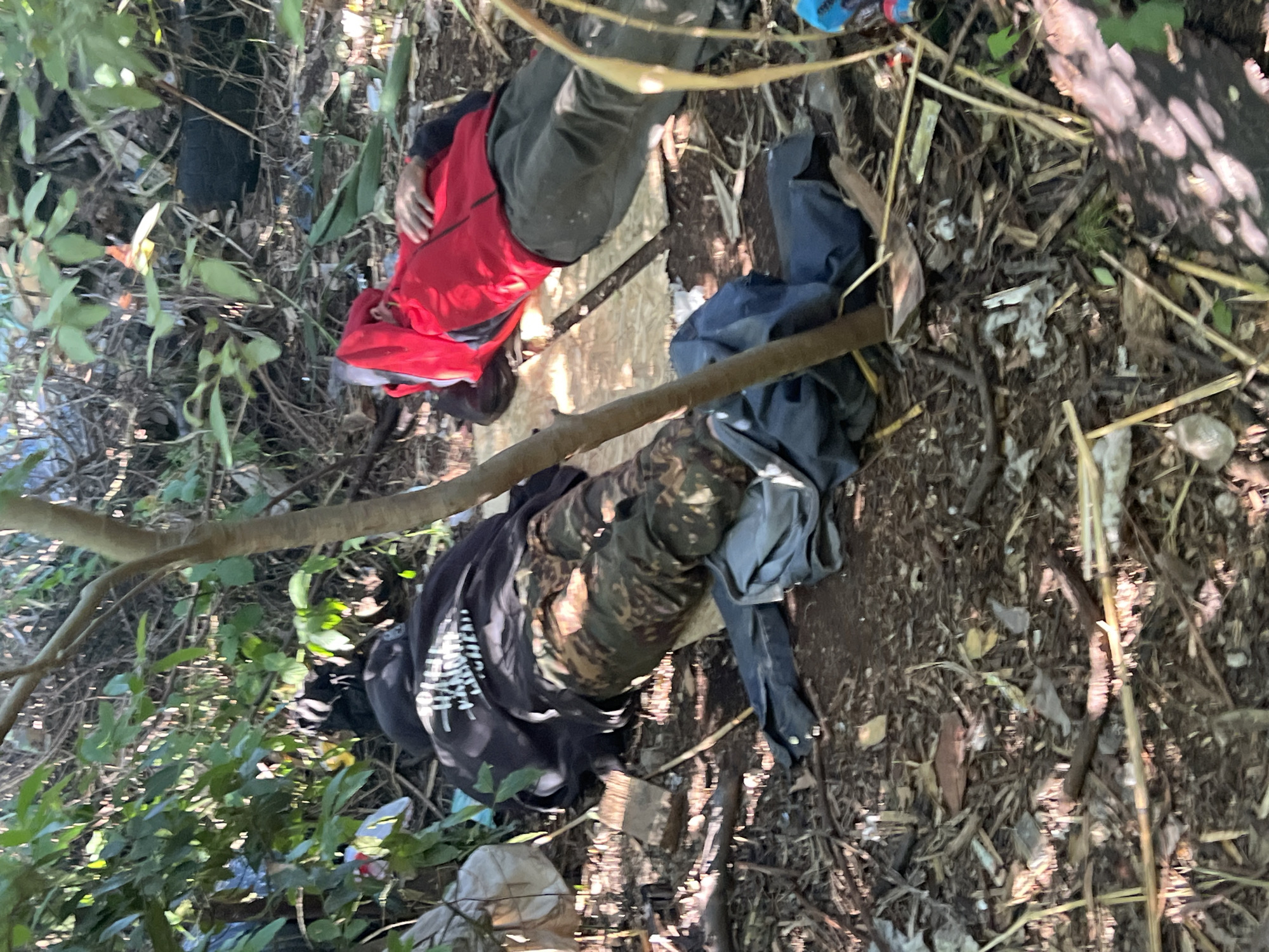 The image shows two individuals lying on the ground in what appears to be a wooded or overgrown area. One person is wearing a red jacket and dark pants, and the other is wearing camouflage pants and a dark shirt with white lettering. The ground is covered with dirt, leaves, and debris, suggesting an outdoor, potentially untidy or natural setting. A piece of wood or plank is visible between the two individuals. The lighting suggests it is daytime, with sunlight filtering through the trees, casting shadows. The overall impression is one of stillness, with the individuals appearing to be resting or possibly incapacitated. The scene lacks any clear indicators of a specific event or activity, beyond the presence of the individuals. There is no visible text that is clearly legible. The context provided indicates the location as Sochi, Russia, which could suggest a natural park, a less maintained area of the city, or a rural outskirts.