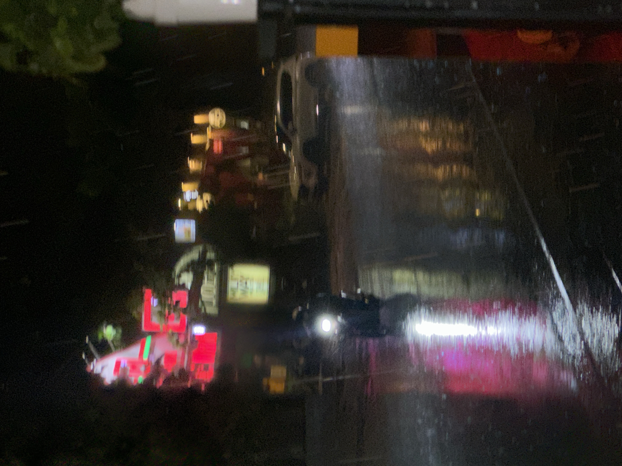 The image depicts a dynamic street scene at night, likely after or during rain, evidenced by the highly reflective and glistening wet road surface. The perspective suggests a view from a slightly elevated position, looking down a thoroughfare.
In the left foreground, a prominent red emergency vehicle, possibly a fire truck or ambulance, stands out with its bright, flashing red and green lights. Its illumination casts strong hues onto the surrounding environment. Moving towards the center of the road, another vehicle, possibly a motorcycle or car, approaches with a powerful, singular headlight that creates an intensely bright, elongated reflection on the wet pavement. Reflections from other vehicles and overhead lights are scattered across the wet asphalt.
The background features numerous blurred lights from buildings and commercial signs, primarily in shades of yellow, red, and white, indicating an urban or commercial area. These lights also produce soft, shimmering reflections on the street. Dark, indistinct shapes of foliage or trees are visible on the far left. The overall mood is one of an active, bustling night, with the wet conditions adding an atmospheric quality to the artificial illumination. No clear, legible text is visible on any signs or objects within the image.
