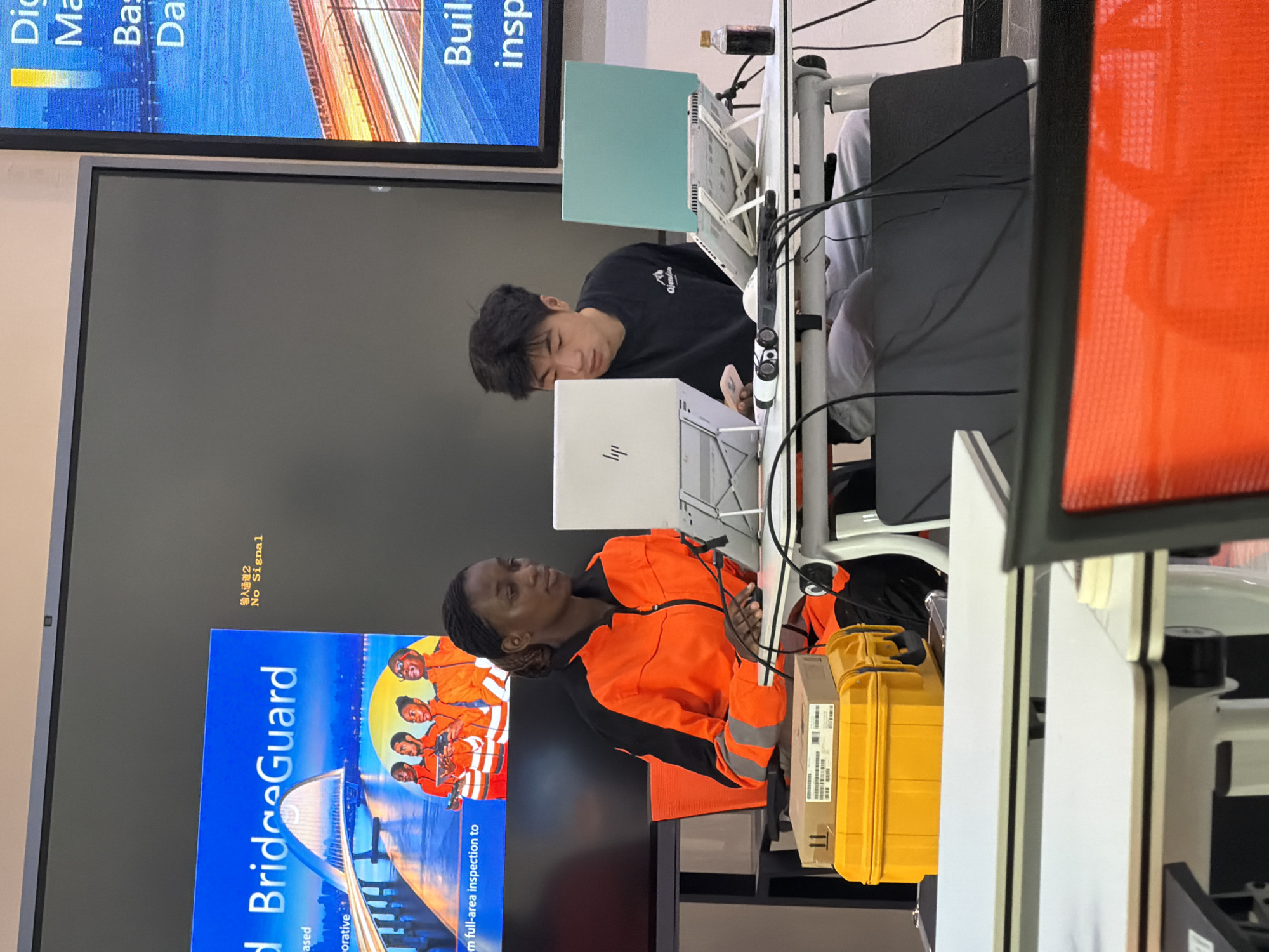 In a well-lit room, two individuals, a woman and a man, are seated at a desk, each with a laptop. The woman, wearing a bright orange safety jacket with reflective stripes, is focused on her screen. To her right, the man, dressed in a black t-shirt, is also engaged with his laptop. The desk is cluttered with various items, including cables, a bottle of dark liquid, and a yellow case with a box on top. Behind them, a large digital screen displays images and text, including the words "BridgeGuard" and "No Signal" in Chinese characters. The overall setting appears to be an office or educational environment, possibly within the city of Jiangjin, China, given the text on the screen. The scene captures a moment of work or study, with the individuals absorbed in their tasks.