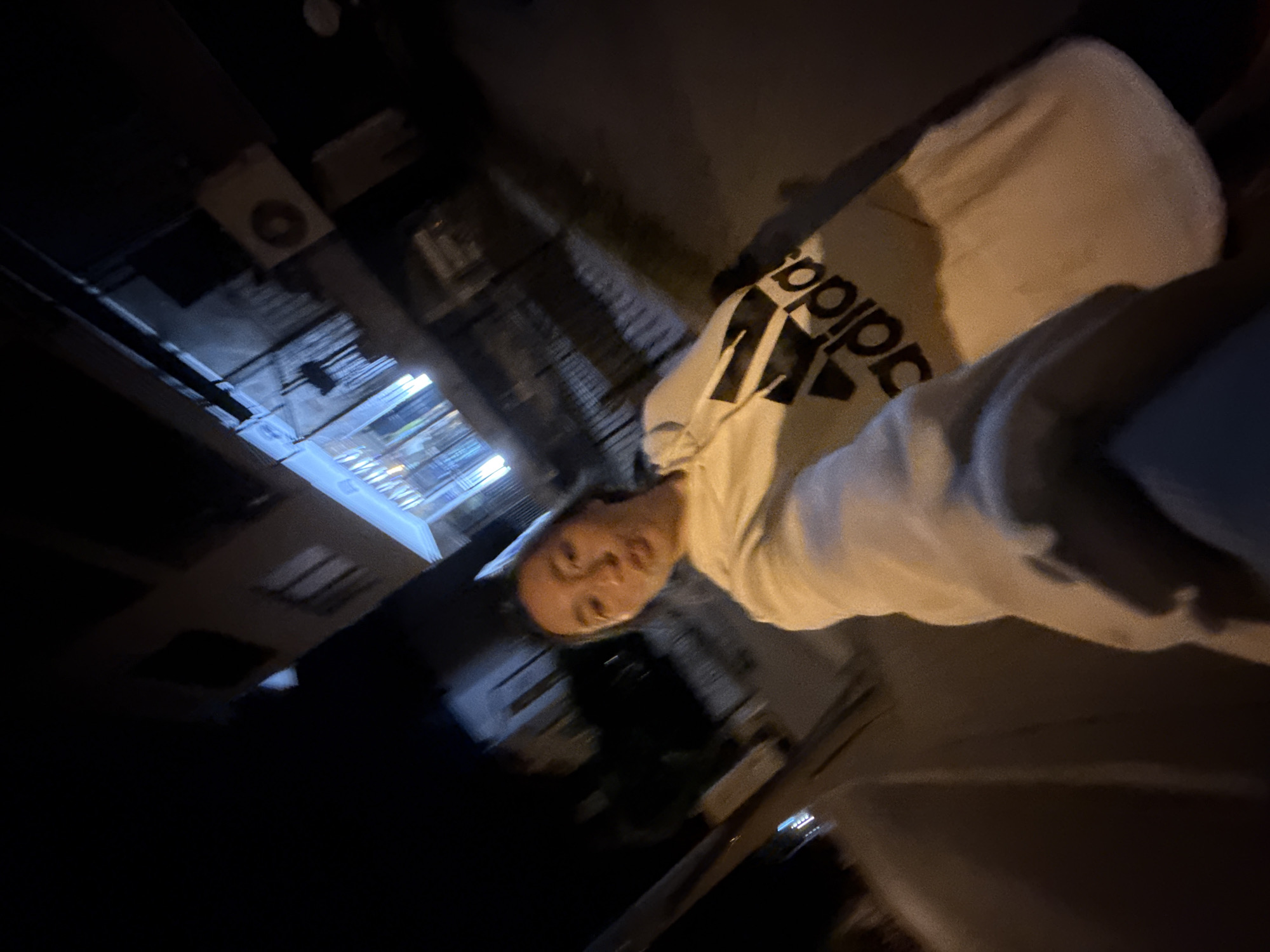 A slightly blurry, low-light, wide-angle selfie taken at night shows a young woman from the chest up. She is wearing a light-colored hooded sweatshirt with a large black Adidas logo on the chest. Her mouth is slightly open, and she appears to be looking towards the camera. The background is dark with blurred lights of a city street, suggesting an urban environment at night. Buildings with illuminated windows are visible in the background. The scene is likely in the city of Sofia, Bulgaria, given the context of previous images.