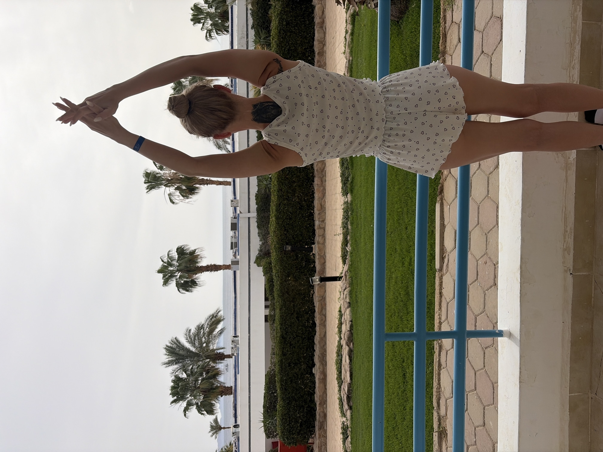 A woman stands on a stone-paved patio with a blue railing overlooking palm trees and the Red Sea, likely at a resort in Sharm El Sheikh, Egypt. She is wearing a white dress with small heart patterns and black socks. Her hair is styled in a bun, and she has tattoos on her neck and upper arm. She is extending her arms overhead, hands clasped together. The background includes a walkway, hedges, grass, and several palm trees against a bright sky.