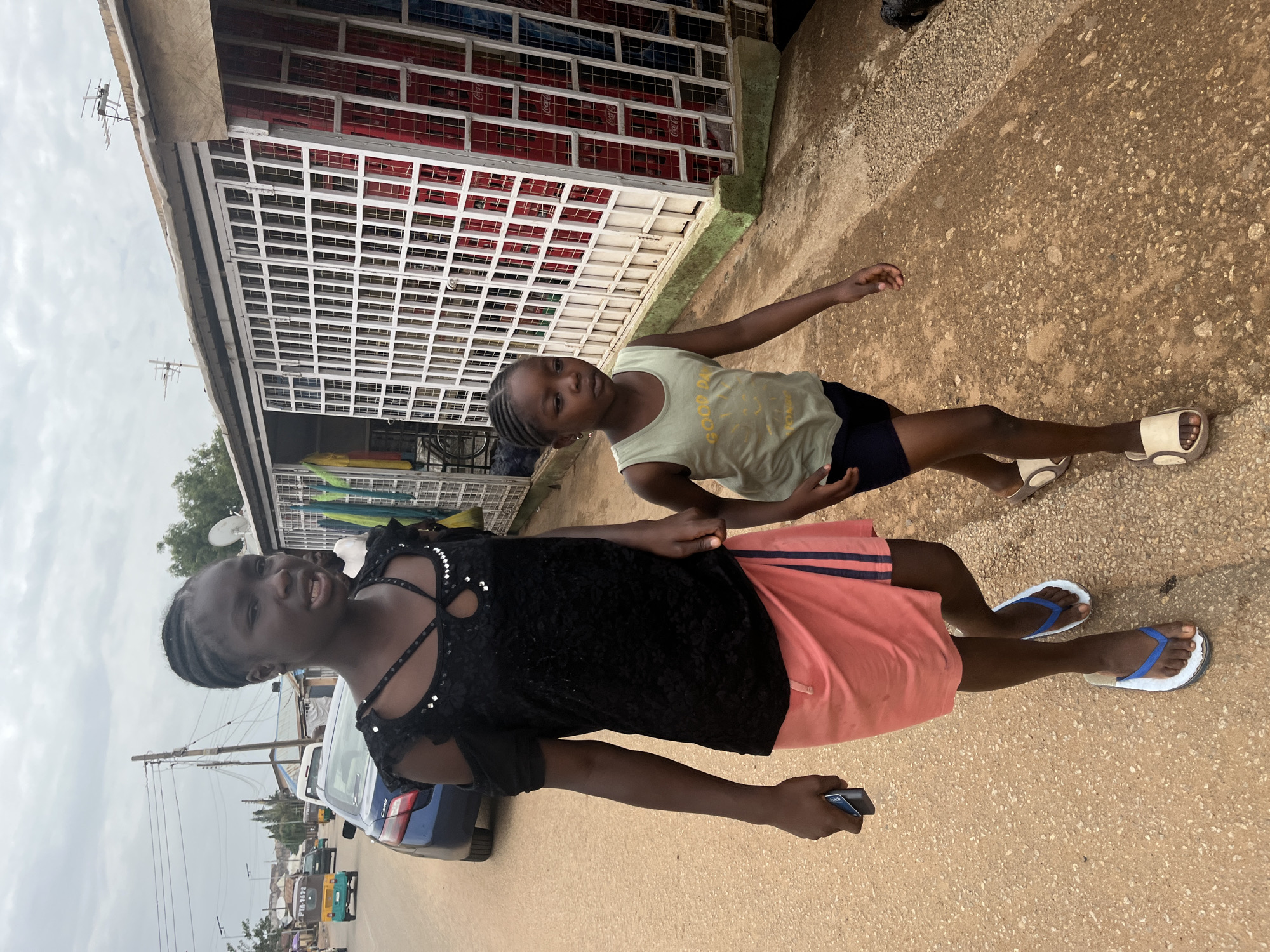 Two young, dark-skinned girls stand in front of a building in Barakin Akawo, Nigeria. The older girl, wearing a black top, peach skirt, and blue flip-flops, holds a cell phone in her right hand. The younger girl, wearing a yellow shirt and black shorts, stands behind the older girl with her arms out. The building behind them has a white grated exterior and is filled with red Coca-Cola crates. The area appears to be a street with cars parked nearby.