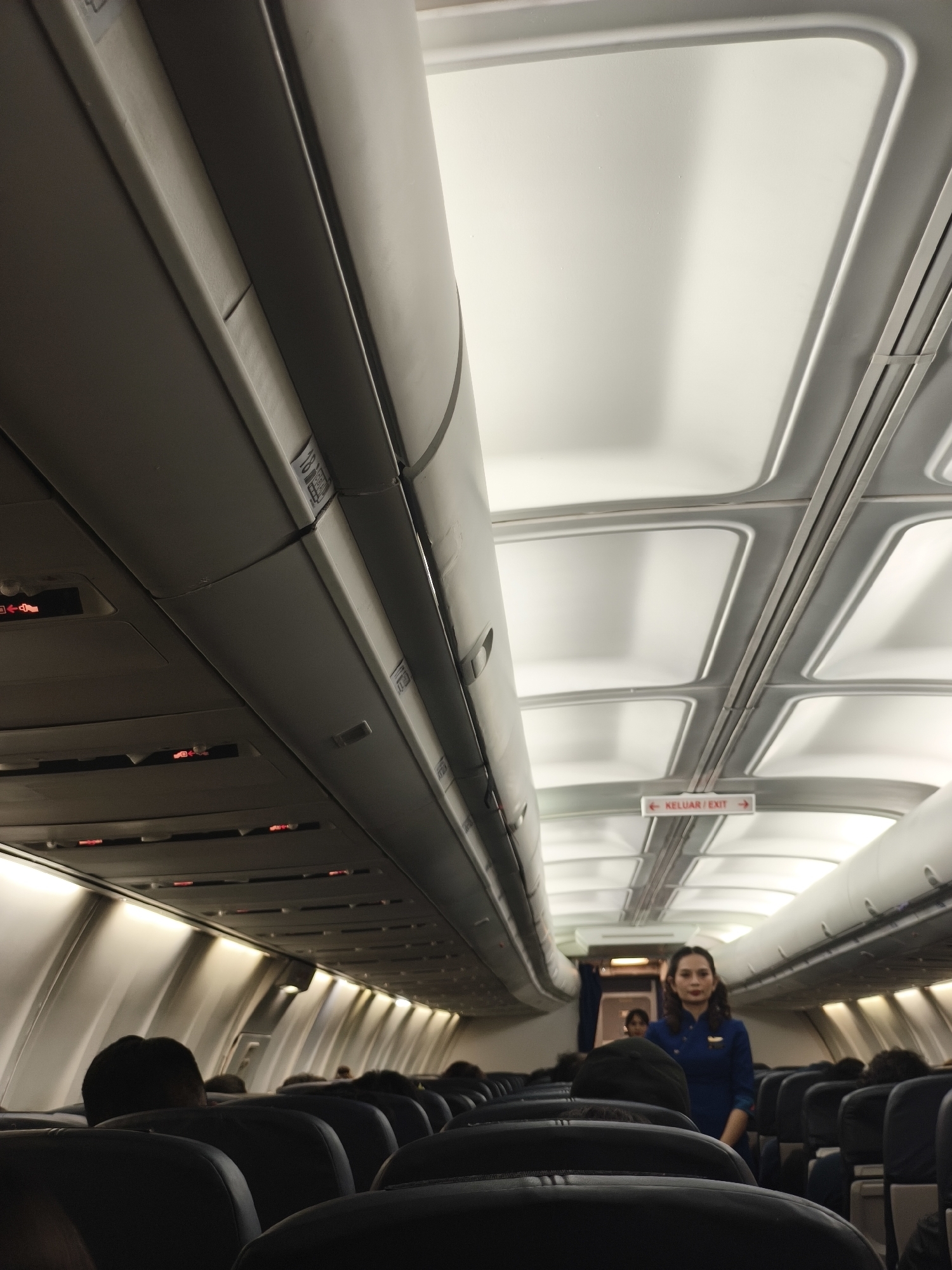 The media file depicts the interior of an airplane cabin. The scene is captured from a low angle, looking upwards towards the ceiling and the aisle. In the foreground, rows of dark, upholstered seats are visible, some with passengers' heads visible over the top. The overhead compartments are closed, with some indicating safety signs and emergency lights.
Further down the aisle, a flight attendant in a blue uniform is walking towards the camera. She has dark hair and is looking directly at the viewer. Another flight attendant is partially visible behind her. Overhead, a sign reads "KELUAR / EXIT" with arrows pointing in both directions, indicating an emergency exit. The cabin's ceiling is lit with bright, diffused lights, creating a clean and modern appearance. The overall impression is of a commercial flight, possibly during daytime given the bright lighting. The presence of the "KELUAR / EXIT" sign in Malay suggests the flight may be in or out of Malaysia or Indonesia, which aligns with the provided location context of Kuta, Indonesia. No specific weather conditions are discernible. The emotions conveyed are neutral, typical of a flight in progress.