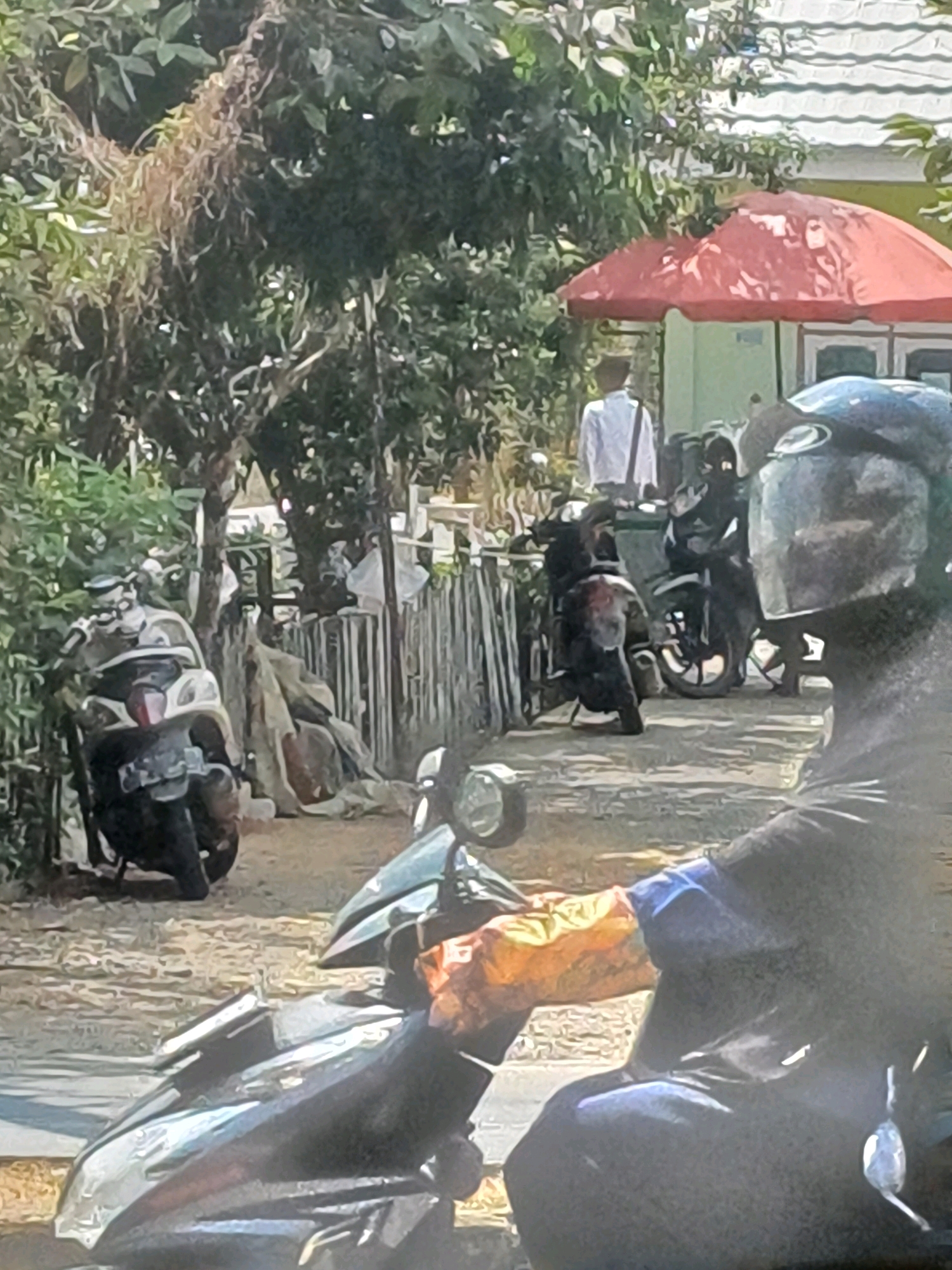 In Pasir Belengkong, Indonesia, an outdoor scene depicts several individuals and motorcycles under a bright, sunny sky, suggesting a warm climate. In the foreground, a person wearing a black shirt and blue trim is seated on a motorcycle, facing away from the camera. They have bright orange gloves on their hands, and a dark helmet with a reflective visor is visible. The motorcycle has a sleek, dark design.
Behind this individual, other motorcycles are parked. One is a light-colored scooter with a black seat and license plate. Another motorcycle, predominantly black, is parked further back, with its front wheel and handlebars visible. A person in a white shirt, carrying a black bag over their shoulder, stands near some greenery and a red umbrella.
The environment features lush green foliage and trees, with some dry, wispy branches visible. A fence, constructed from vertical wooden planks, runs through the midground. Buildings with white roofs are visible in the background, suggesting a residential or commercial area. The ground is unpaved, appearing to be dirt or gravel. The overall impression is that of a casual, everyday scene in a tropical or subtropical setting.