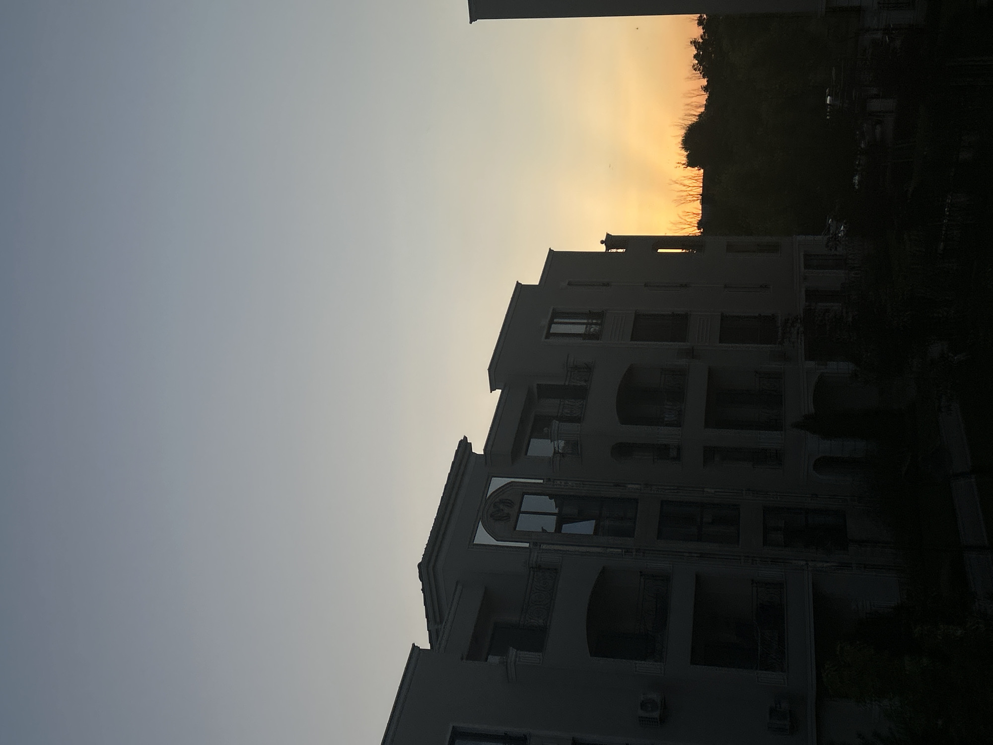 The image depicts a residential building in Plovdiv, Bulgaria, at sunset. The sky is a gradient of pale blue to orange. The building is modern with balconies and ornate window frames. Silhouetted trees and a second building are visible in the background. The lighting suggests it is dusk.