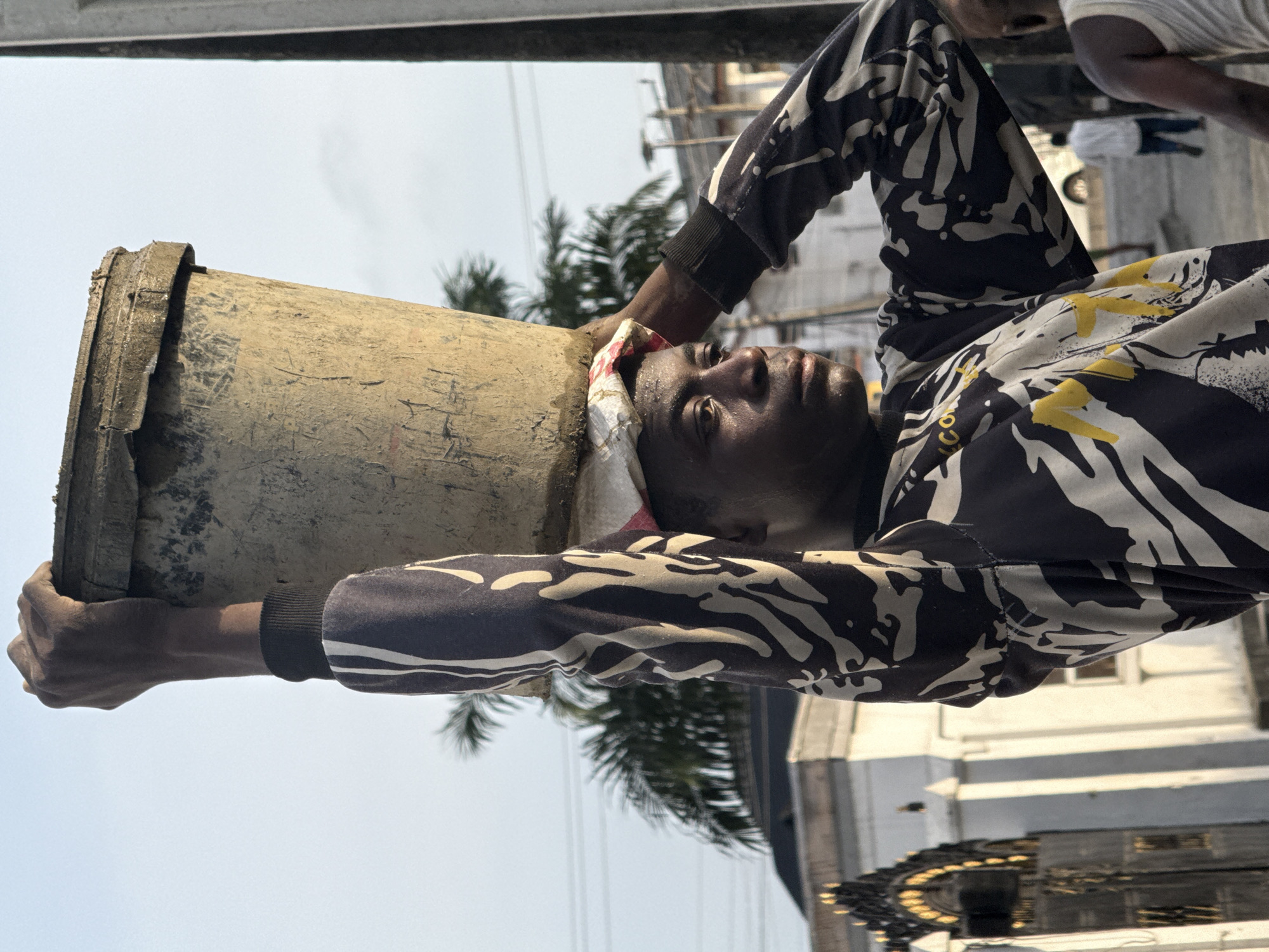 A man is carrying a large, tan, cylindrical bucket on his head in Ado, Nigeria. He wears a patterned black and white long-sleeved shirt and a white cloth under the bucket for support. The bucket appears to be made of a rough, textured material, possibly cement or plaster. Palm trees and buildings are visible in the background. The sky is a hazy pale blue. The man is looking upwards with a slight grimace. He grips the bucket with both hands.