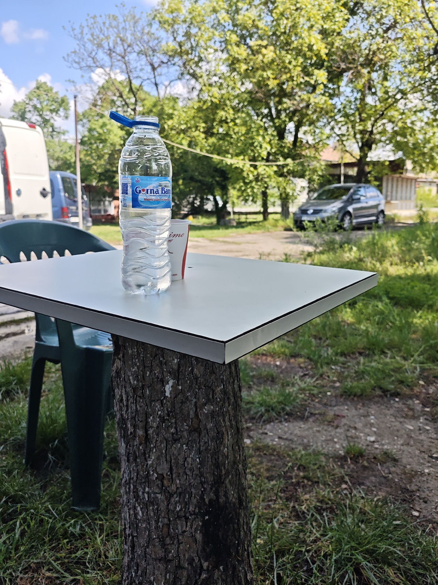 The image depicts an outdoor setting featuring a white rectangular table with a tree stump serving as its base. On the table, a clear plastic water bottle with a blue label is prominently displayed. The label on the bottle reads "Gorna Banya" and "Mineral water for your daily", and the bottle has a blue flip-top cap. Behind the bottle, a white and red paper cup, partially obscured, shows the text "imo".
To the left of the table, a green plastic chair is visible. The foreground consists of grassy ground and some dirt patches. In the background, under a bright blue sky with scattered white clouds, a lush canopy of green trees dominates the landscape. Various vehicles are present in the mid-ground: a white van and a blue van are seen on the left, and a gray car is parked further in the distance on the right, partially hidden by foliage. The ground in the background appears to be a mix of dirt, grass, and possibly a gravel path.
The scene suggests a casual outdoor location, perhaps a rest stop or a simple outdoor seating area in a rural or semi-rural environment. It is daytime, with clear, sunny weather. No people are visible, lending a peaceful and tranquil atmosphere to the image.