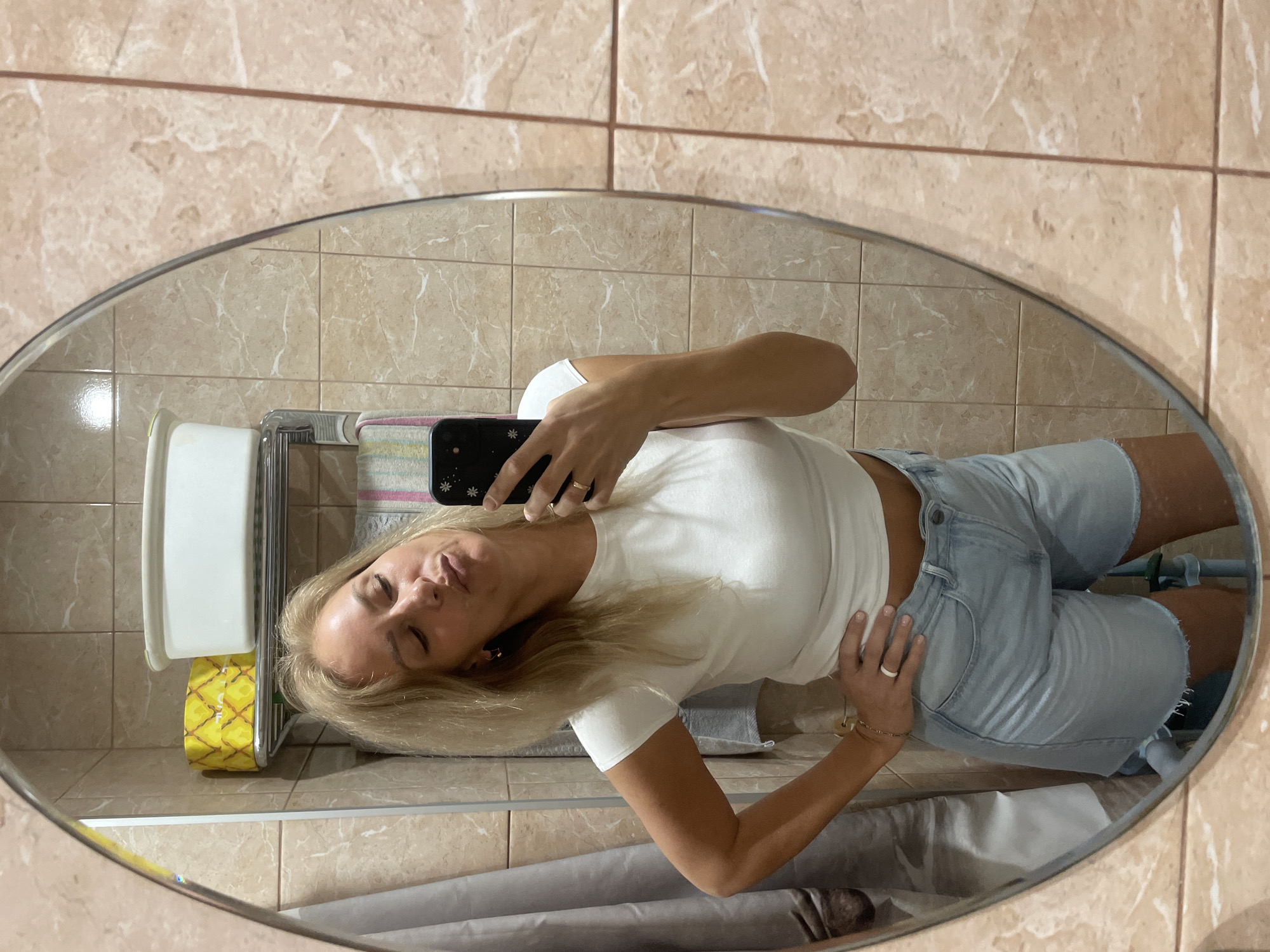 A woman with long blonde hair is taking a selfie in a bathroom mirror. She is wearing a white t-shirt and light blue denim shorts. The bathroom has tan-colored tiled walls. A toilet paper holder with yellow toilet paper is visible next to the mirror. A towel rack is situated above what appears to be a gray curtain or fabric. The overall scene is indoors. The location in the city of Sochi, Russia, cannot be determined from the image.