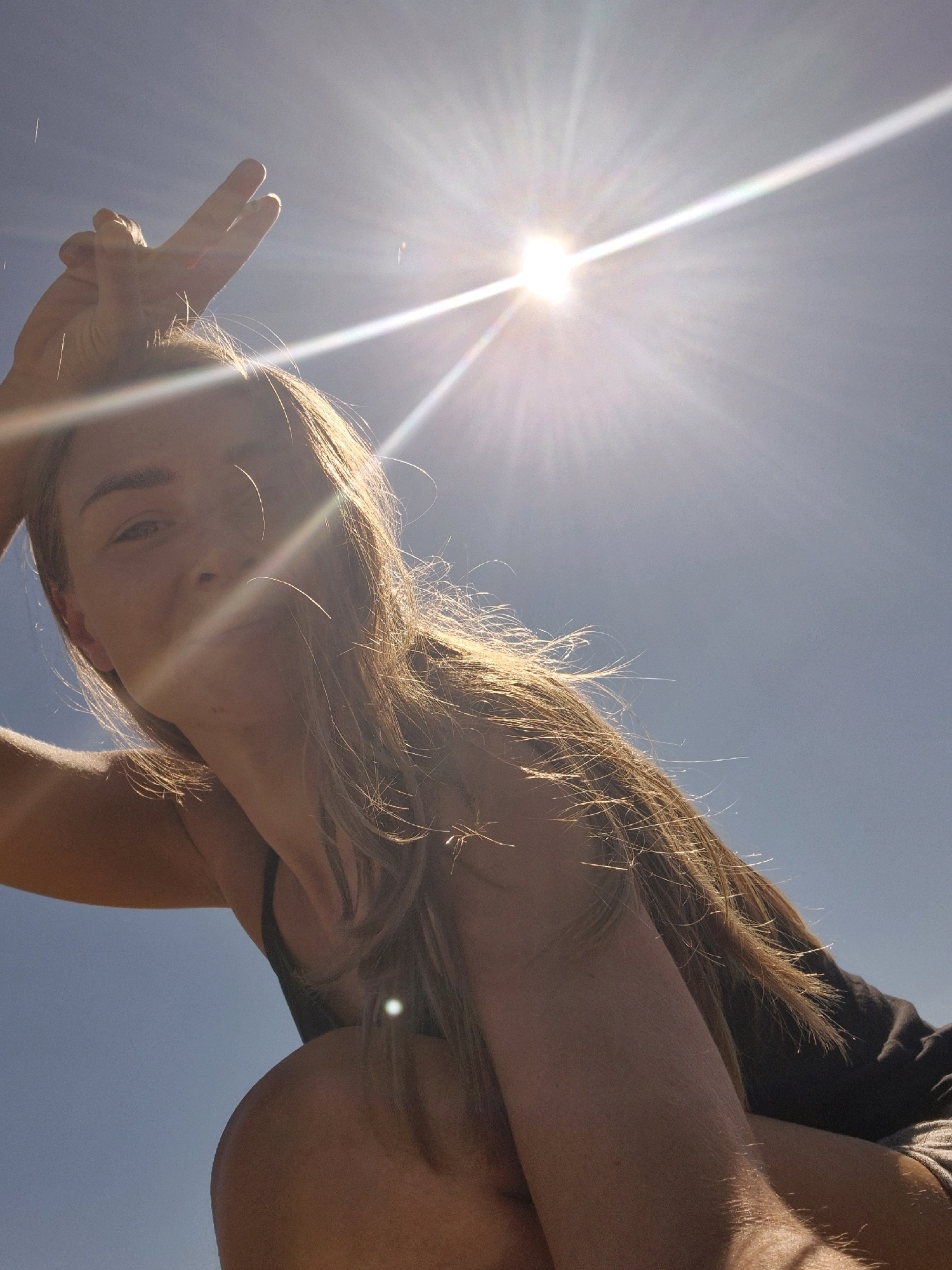 A low-angle, outdoor photograph shows a woman looking up at the sun. The woman has long, blonde hair and is wearing a black tank top. She is holding up her right hand, with her index and middle fingers extended in a peace sign. The sun is a bright, direct light source, creating lens flare and strong highlights on the woman's hair and skin. The background is a clear blue sky. The image appears to have been taken in the city of Zagreb, Croatia, possibly on a sunny day.