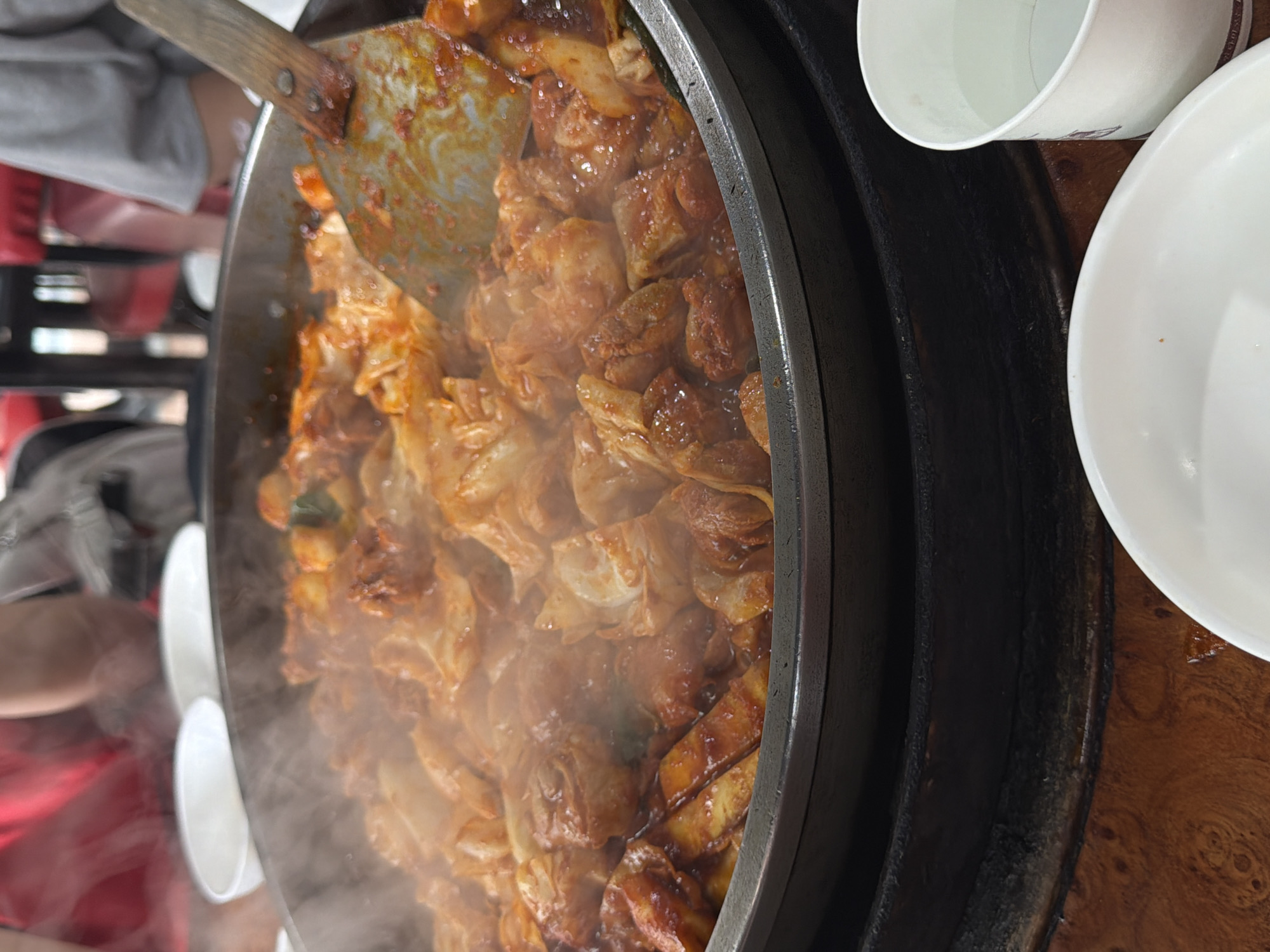 A close-up, overhead shot shows a large, round, black metal pan filled with steaming, vibrant red and orange Korean stir-fried chicken, known as Dakgalbi. The dish is a mixture of chopped chicken, cabbage, and what appear to be rice cakes or sweet potato slices, all coated in a spicy sauce. A metal spatula, stained with the same sauce, is embedded in the food, suggesting it is actively being cooked or served. Wisps of steam rise from the pan, indicating the heat of the dish. In the background, out of focus, are blurry figures of people wearing casual clothing, seated at a table in what appears to be a restaurant setting. The table is wooden, with a visible grain pattern. To the right of the pan, a white paper cup containing a clear liquid, likely water, is partially visible. To the left and in the foreground, two white ceramic bowls, one partially filled with food, are also present. The overall scene suggests a communal dining experience in Chuncheon-si, South Korea, a city renowned for its Dakgalbi.