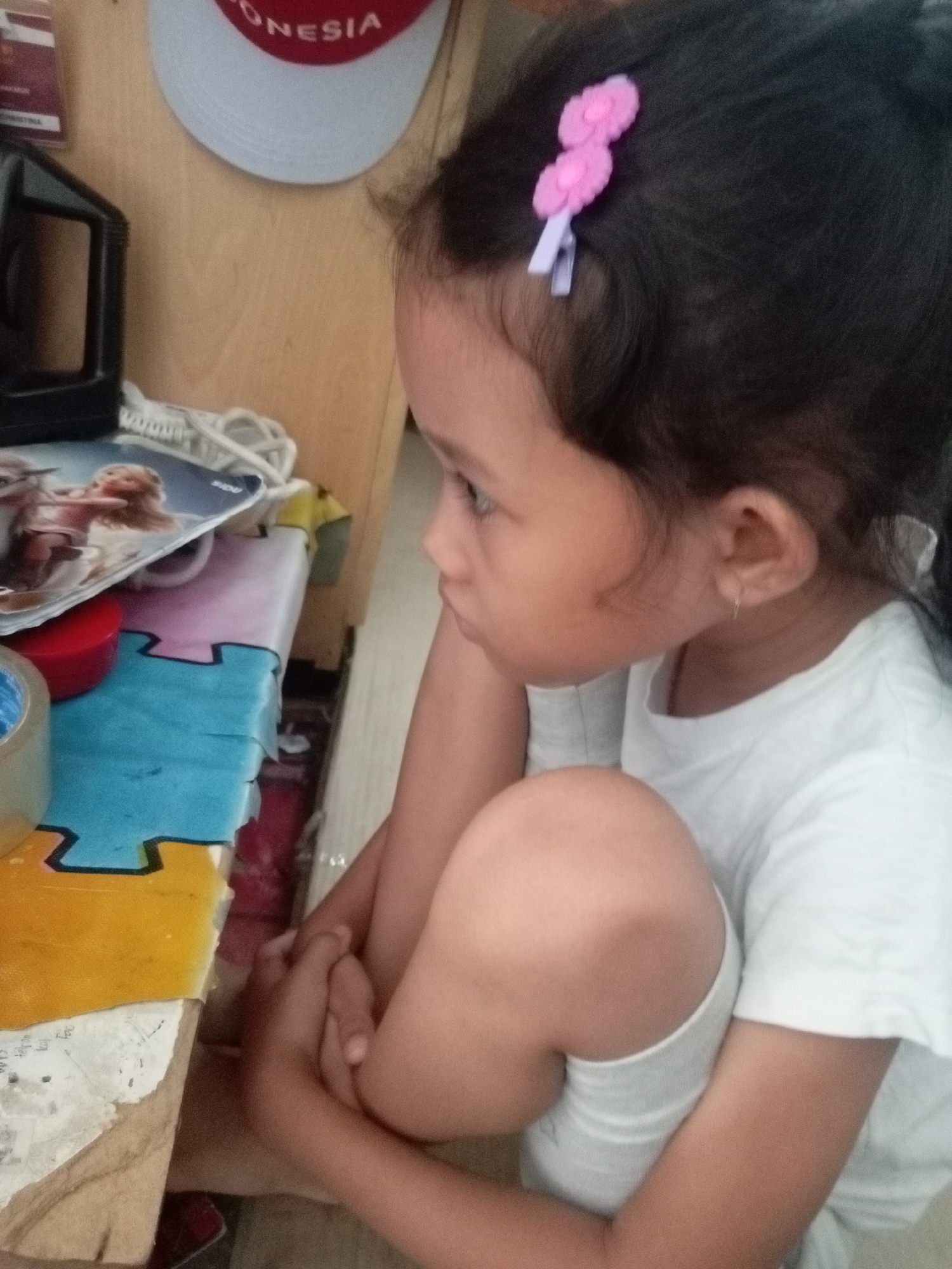 The image shows a young girl in a white t-shirt sitting on a floor, with her knees pulled up to her chest. She has dark hair with a pink flower clip. She is looking to her left, with her head resting on her arm. In the background, there is a wooden shelf with various items on it, including a black object and what appears to be a book or magazine with images of cartoon characters. There is also a blue and yellow puzzle mat on the floor. The setting appears to be a home environment. No specific location in Batam, Indonesia is identifiable from the image.