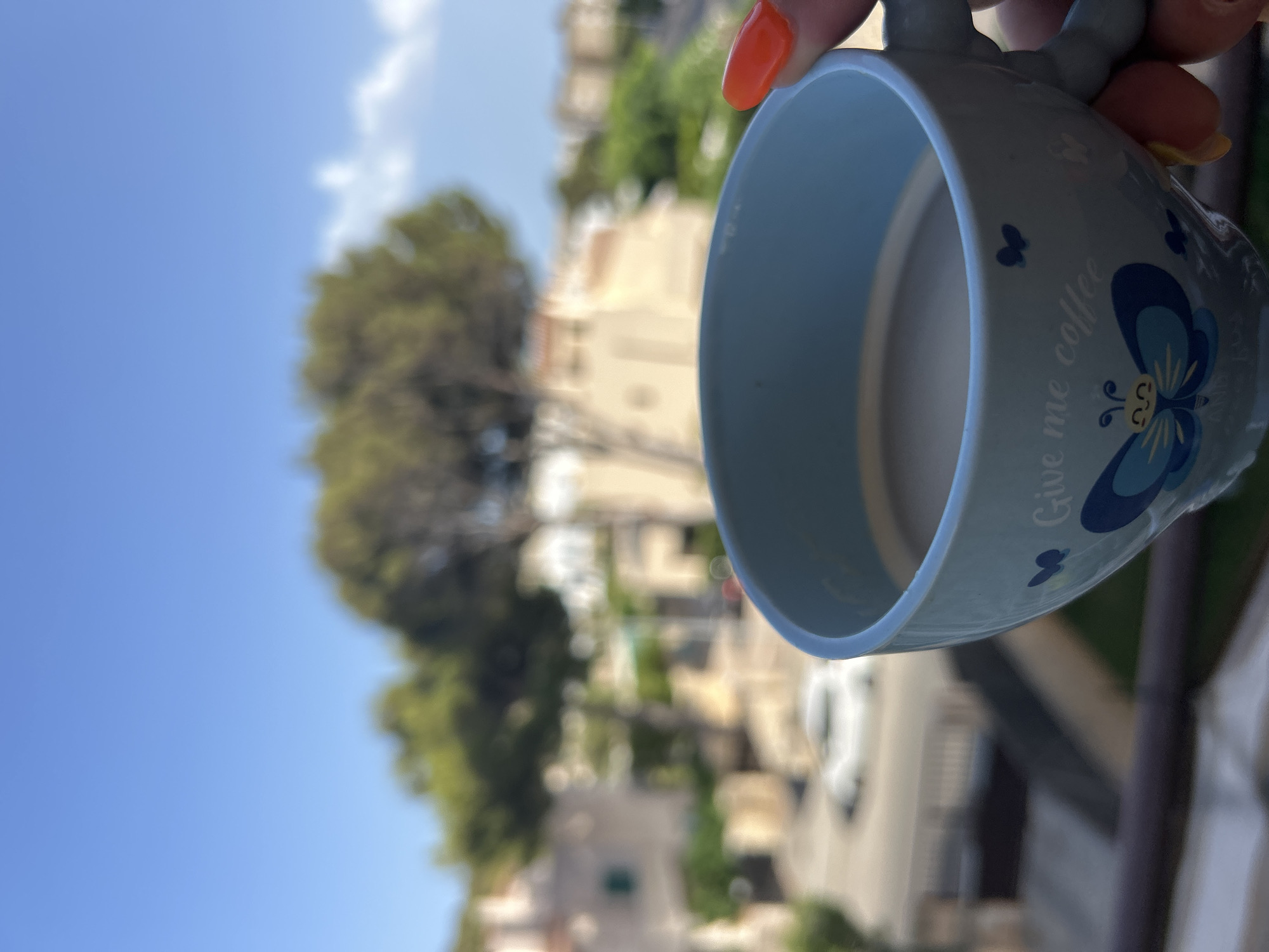 A close-up, shallow depth-of-field shot features a light blue ceramic mug filled with coffee, held by a hand with bright orange nail polish. The mug has a butterfly design and the text "Give me coffee" printed on it. In the blurred background, a sunny day is depicted with clear blue skies, some white clouds, green trees, and light-colored buildings typical of Capdepera, Spain. A car is visible in the distance on a street.