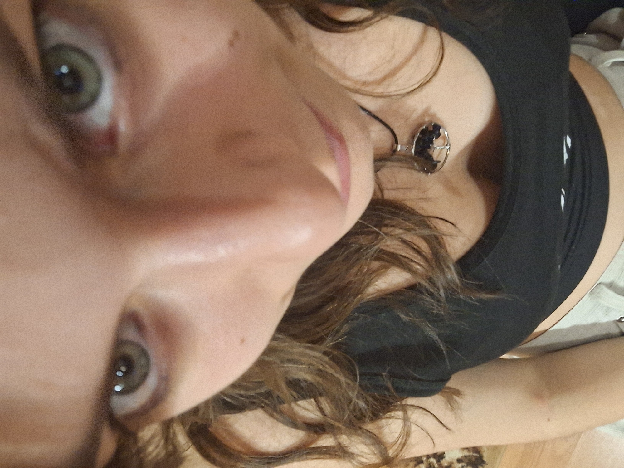 A close-up, angled selfie shows a woman with green eyes, brown, curly hair, and fair skin. She wears a black top and a necklace with a silver pendant. The background is blurry and indistinct, suggesting an interior location. The image does not contain information to determine the location in Panagyurishte, Bulgaria.