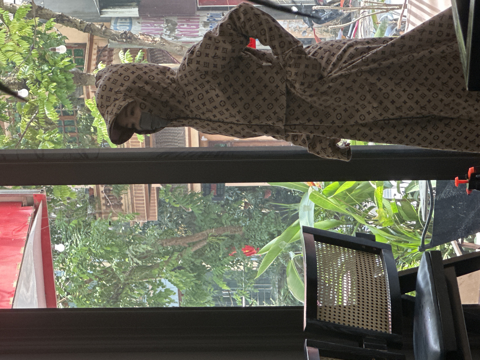 A person is depicted standing upright, framed within the lower portion of a window, looking out onto a vibrant outdoor scene. The individual is wearing a full-body garment, possibly a hooded jumpsuit or coordinated top and pants, adorned with a repeating pattern resembling the Louis Vuitton monogram, featuring stylized "LV" initials, stars, and flowers. A grey face mask covers the lower half of the person's face.
Through the clear window, lush green foliage from trees dominates the view, with some light bulbs hanging within the branches. Buildings with traditional-style wooden shutters and awnings are visible behind the greenery, suggesting an urban or semi-urban environment. A bright red flower is seen peeking through the leaves. On the left side of the window, a portion of a red structure or awning is visible, and on the right, a dark-framed chair with a woven cane back is partially in view, indicating an indoor setting.
The scene is brightly lit by natural light, suggesting it is daytime. The overall impression is of someone looking out from an interior space into a lively outdoor environment. Visible text through the window, albeit partially obscured and faded, includes parts of signs that appear to read "HOTE" (likely part of "HOTEL") and "RANG" (consistent with the location context of Phố Ràng, Vietnam), along with "VN" and "PHI".