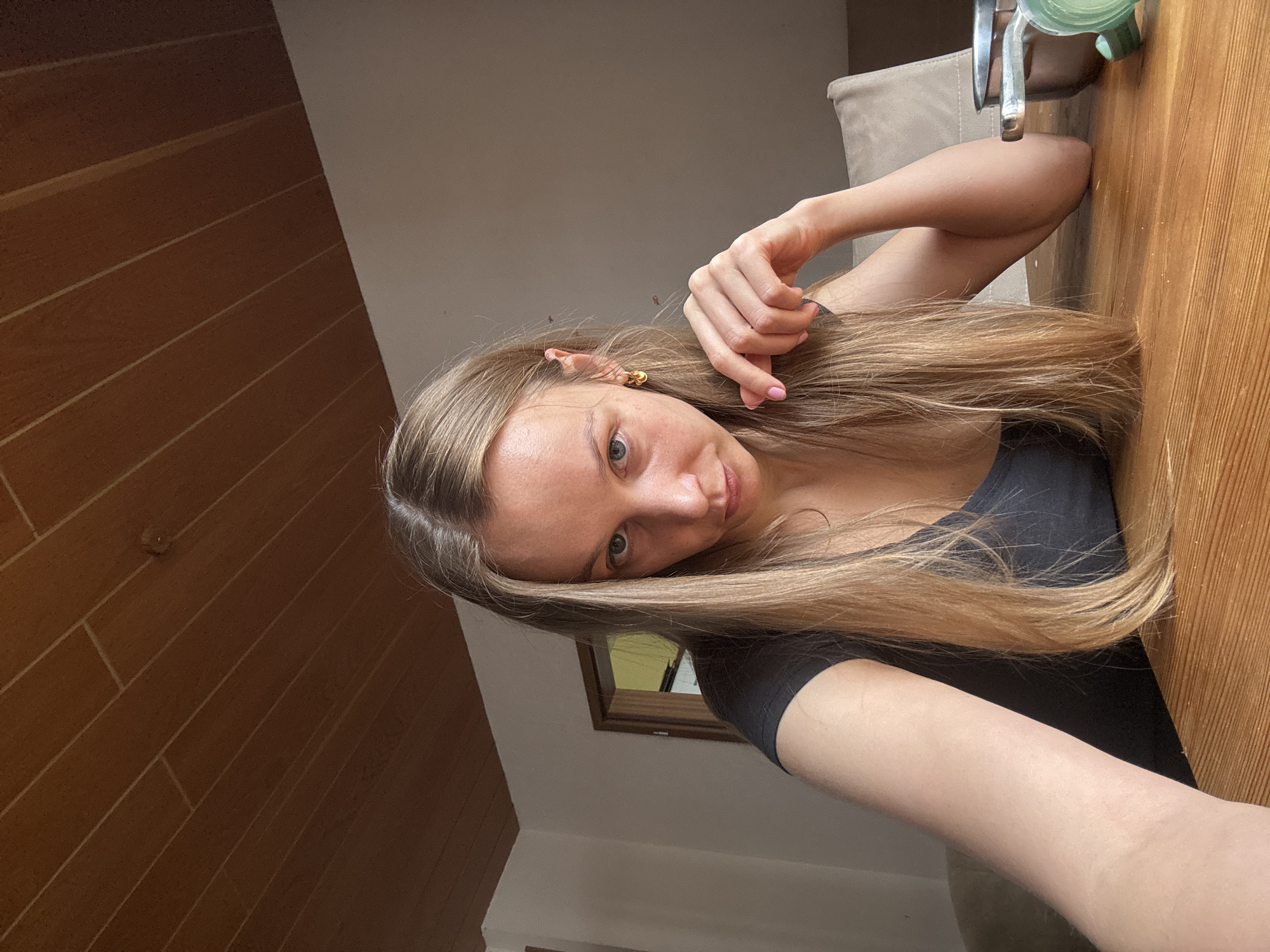 A young woman with long, blonde hair is seated at a wooden table, looking directly at the camera. She is wearing a dark top. Her right hand is resting near her chin, with her fingers gently touching her hair. The background shows a wall and a doorway. A metallic bowl is visible on the table to her right. The ceiling above is made of wood paneling. The scene appears to be indoors, possibly in a home setting in Überherrn, Germany, given the context provided.