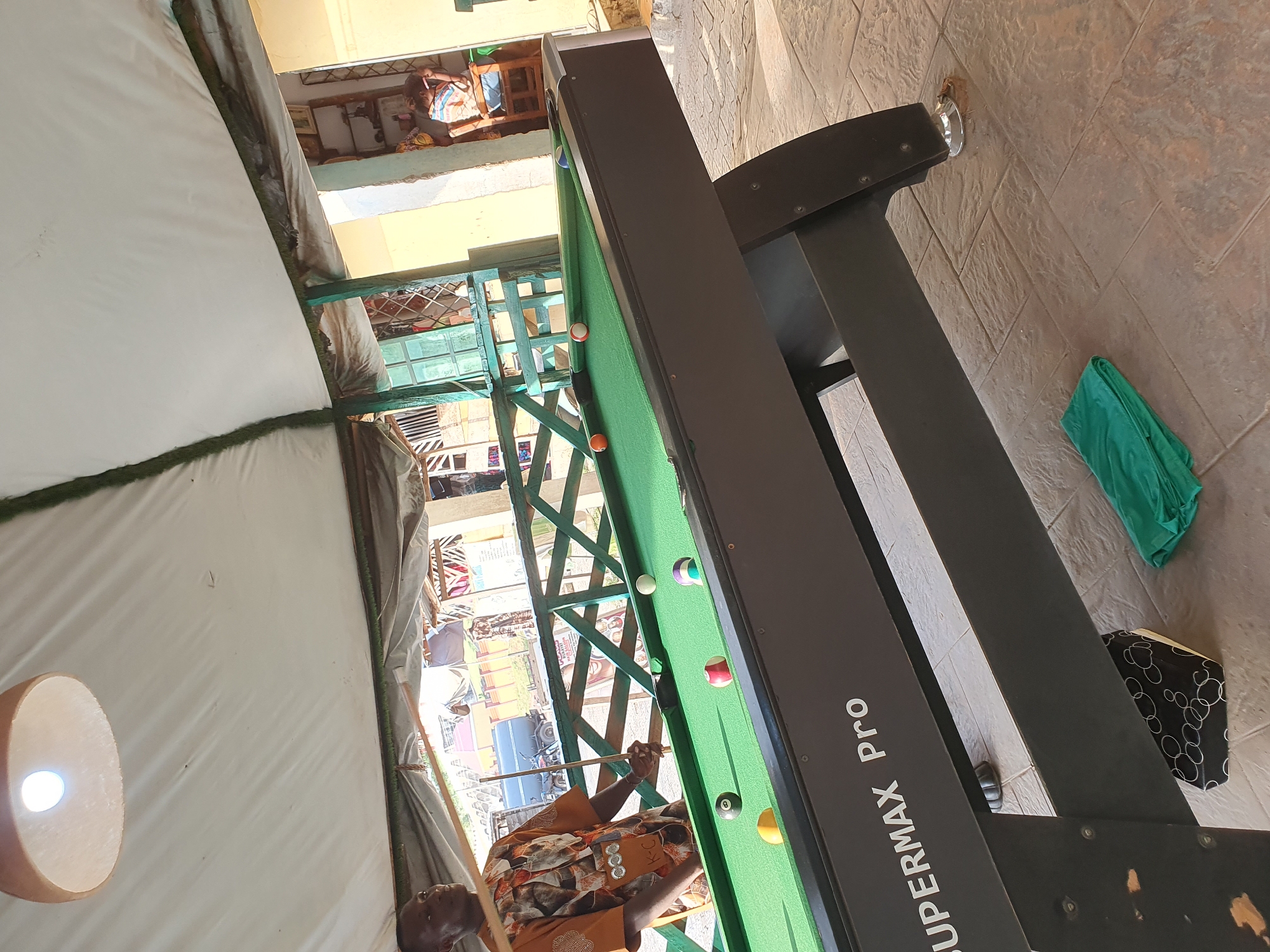 In Lokoja, Nigeria, a man stands by a green pool table with several balls scattered across its surface. The man, wearing an orange and white patterned shirt, holds a pool cue. The pool table, labeled "SUPERMAX Pro," is positioned outdoors under a white canopy supported by green posts. In the background, buildings and a truck are visible. A folded green cloth rests on the tiled floor near the pool table's leg.