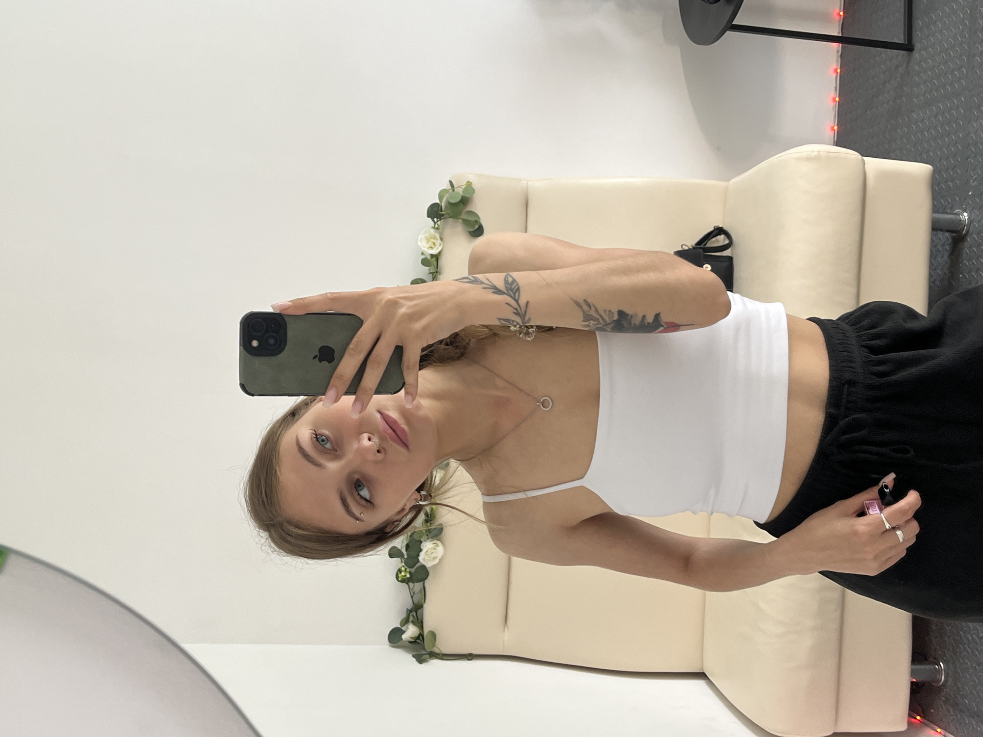 A young woman is captured in a mirror selfie, standing indoors. She has light brown hair, blue eyes, and visible piercings on her eyebrow and septum. She is wearing a white spaghetti-strap crop top and black sweatpants or joggers. Tattoos are visible on both of her arms: a leafy branch design on her right forearm and a more abstract, dark design on her left upper arm. She is also wearing a silver necklace with a circular pendant and several rings on her fingers, including a pink one. Her fingernails are painted a light pink or nude color.
She holds a dark green iPhone with an Apple logo clearly visible on its case, pointing it towards the mirror to take the photo.
The background features a light-colored, possibly cream or beige, sofa. White artificial roses and green foliage are draped decoratively over the back of the sofa on the left side. A black bag is placed on the sofa next to her. The room has white walls and a dark grey, possibly textured or rubberized, floor. In the upper right background, red string lights are visible along a dark metal structure. A curved edge of the mirror is visible in the bottom left corner of the frame. There is no visible text in the image apart from the Apple logo on the phone.