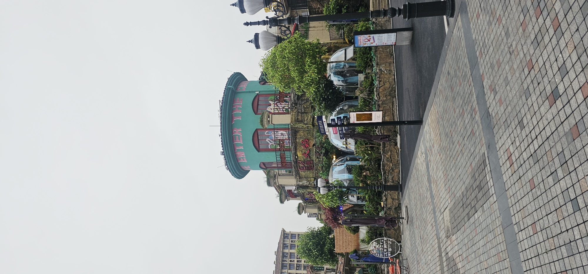 The photograph depicts a street scene in Chongqing High-tech Zone, China, characterized by distinctive architectural elements and a modern urban environment.
The most striking feature is a multi-story building, identifiable by its turquoise exterior and red window frames. The phrase "ENTER THE GATE" is vertically written on the building's facade in red letters. Graffiti is visible on some parts of the building. The building is surrounded by dense foliage and decorative elements, including planters and ornate metal railings.
Adjacent to the building is a paved pedestrian walkway made of small, multi-colored square tiles in a grid pattern. Next to the walkway is a narrow street, with vehicles visible in the background. Tall black lampposts are spaced along the edge of the street. Further down the street, there are transparent dome-shaped structures, likely seating areas or decorative installations.
In the distance, additional buildings are visible, suggesting a bustling urban setting. The sky is overcast, creating a soft, diffused light.
