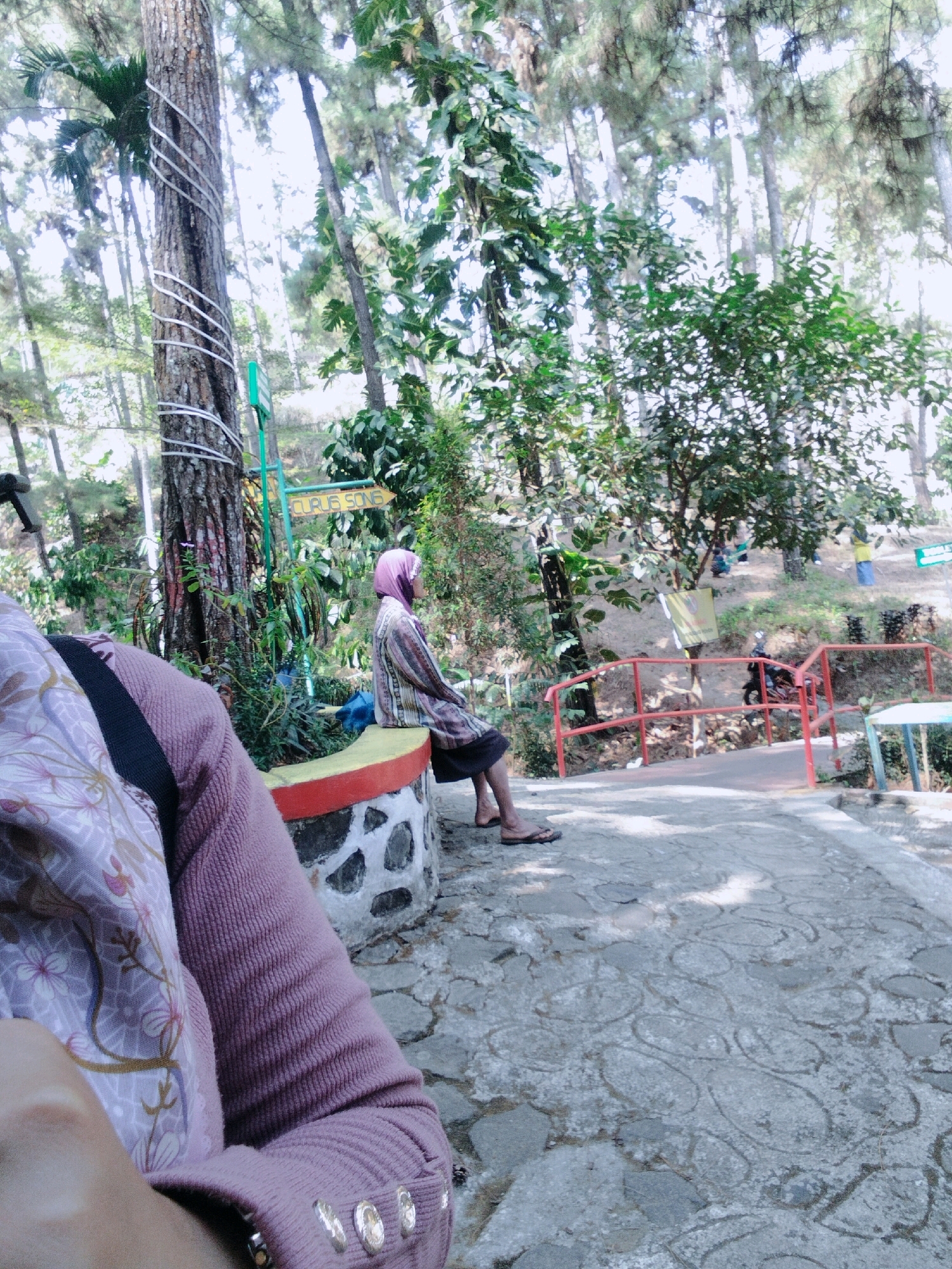 In the city of Kaliwedi, Indonesia, a medium-shot outdoor photograph captures a serene park scene. In the foreground, the left arm of a person wearing a textured purple sweater with floral patterned fabric is visible, partially obscuring the view. The person is sitting on a stone-and-concrete bench with a yellow and red trim. To the right, a woman in a headscarf and a patterned shirt is seated on the same bench, facing away from the camera. Her legs are crossed, and she is wearing sandals. The background reveals a pathway made of patterned concrete stones, leading through a lush, green environment with numerous trees and foliage. A red railing delineates a slightly elevated area to the right, where a motorcycle is parked. Two signs are visible, one green and one yellow with text. The overall atmosphere suggests a tranquil spot for relaxation and recreation within a natural setting.