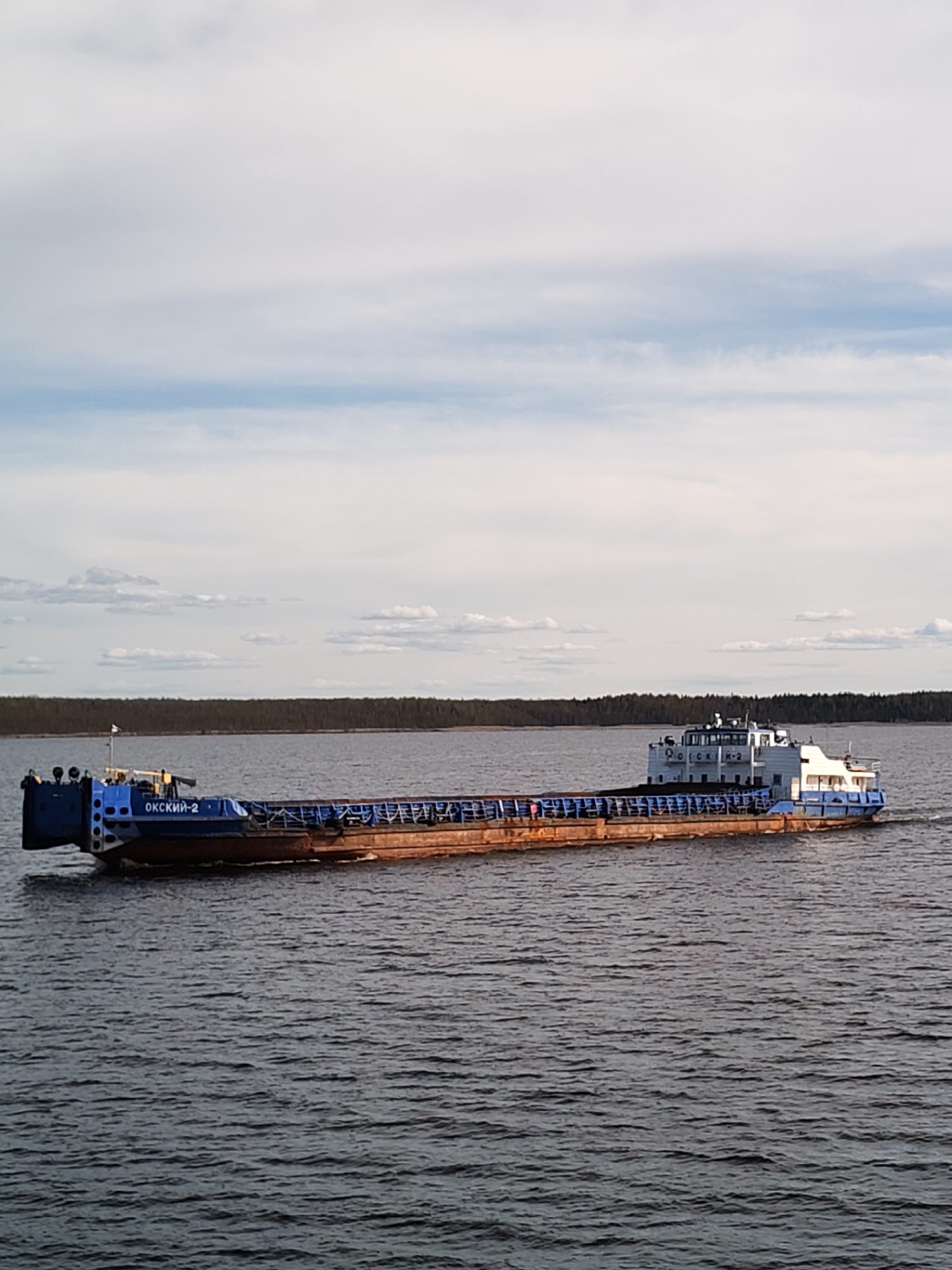 A barge labeled "OKCKNN-2" is sailing on a body of water in Gorushka, Russia. The barge is primarily rust-colored with blue sections at the front and rear. A cabin-like structure is visible at the rear. The water is choppy. A strip of trees lines the horizon in the background, and a cloudy sky occupies the upper half of the image.