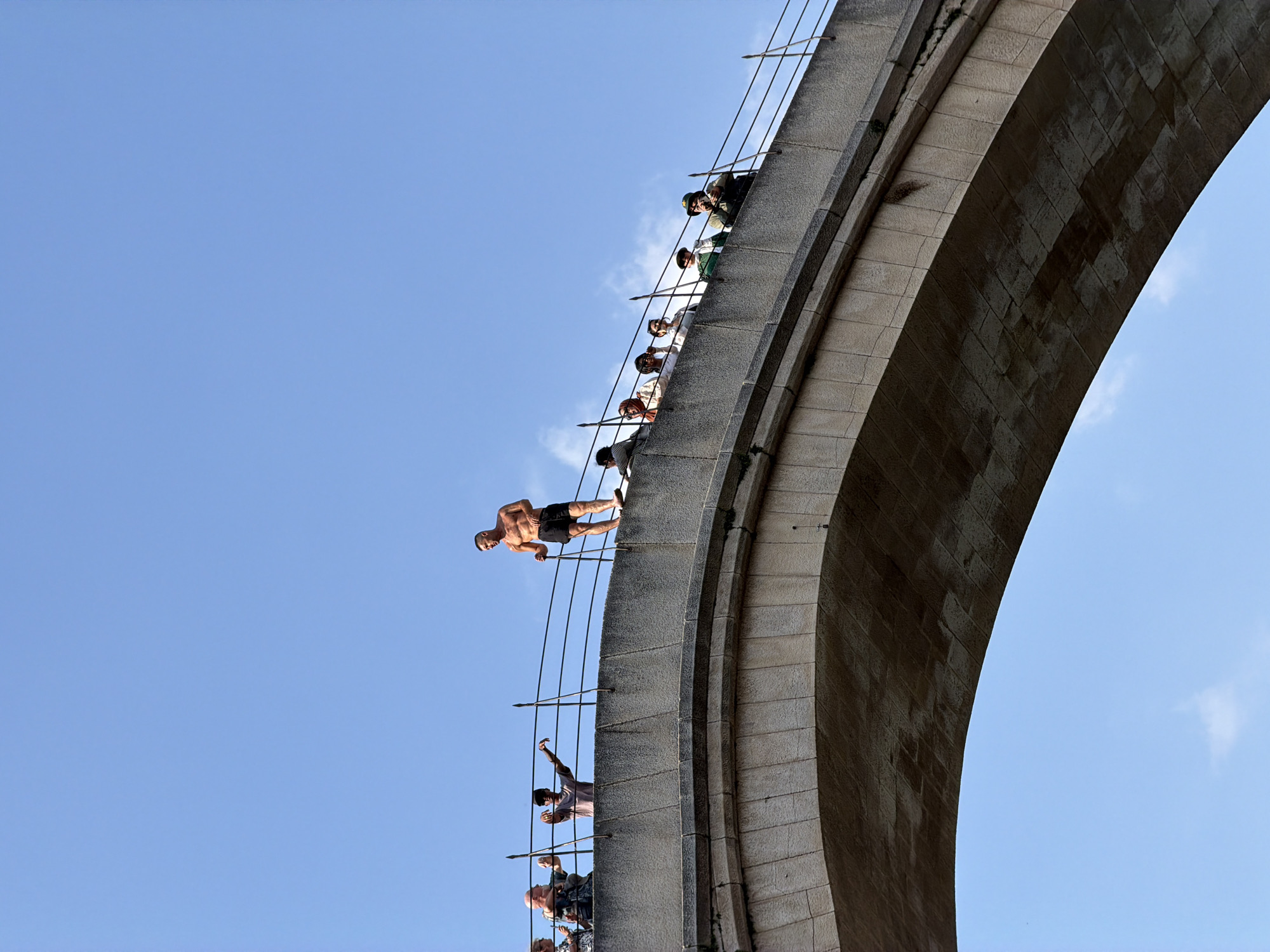A shirtless man stands on a stone bridge, holding a stick in his right hand. He wears black shorts. He is on the edge of the bridge, facing left. Several people are visible on the bridge, behind him, looking in the same direction. The bridge has stone arches and is lined with a railing. The sky is clear and blue. This is likely the Stari Most (Old Bridge) in Mostar, Bosnia and Herzegovina, a famous diving spot.