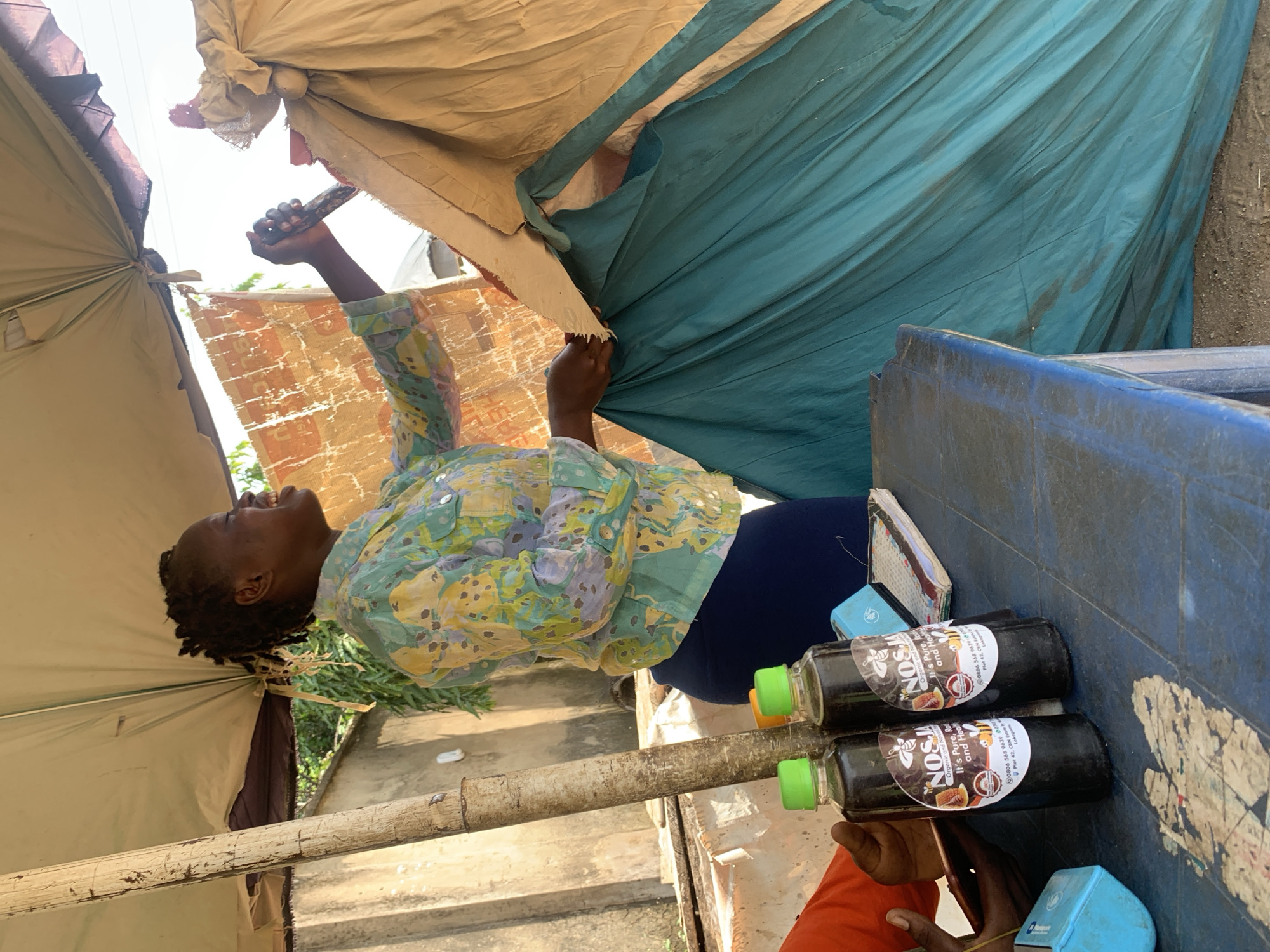 In Abuja, Nigeria, a woman with dark braided hair, wearing a colorful patterned shirt and a dark blue skirt, is seen laughing and holding a stick. She is standing under a beige tarp supported by a wooden pole. In front of her, on a blue plastic table, are two bottles of what appears to be honey, labeled "NOSJA Organic and Natural". The bottles have circular labels featuring an illustration of a honeycomb with bees and text. To the left of the bottles are other objects on the table, including what looks like a blue credit card reader and a notebook. The background consists of a concrete wall and some foliage. A large teal-colored tarp hangs behind the woman, obscuring part of the background. The overall scene suggests an outdoor market or roadside stall.