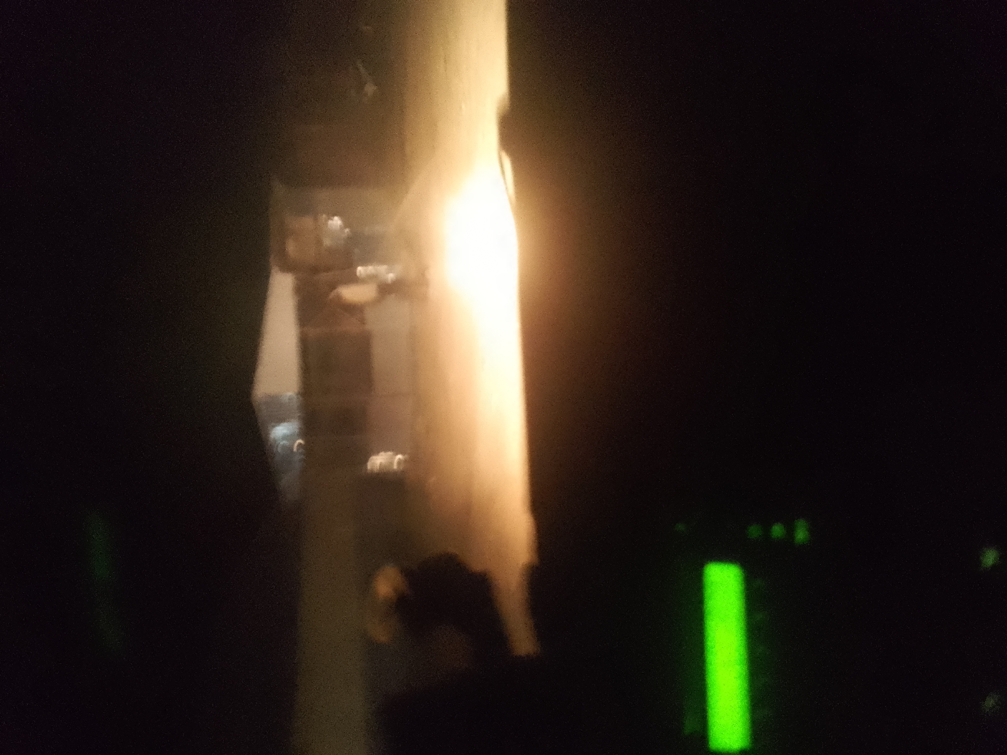 The image is a low-light, out-of-focus photograph taken at night, likely from inside a vehicle. In the center of the frame, a bright, vertical light source illuminates a textured, light-colored surface, possibly a wall or a column. To the left of this light source, a blurred reflection or object can be seen, with hints of white light. The right side of the image is predominantly dark, with a faint green light in the lower right quadrant. The overall scene suggests an urban environment at night, with artificial lighting being the primary visual element. The exact location is unknown.
