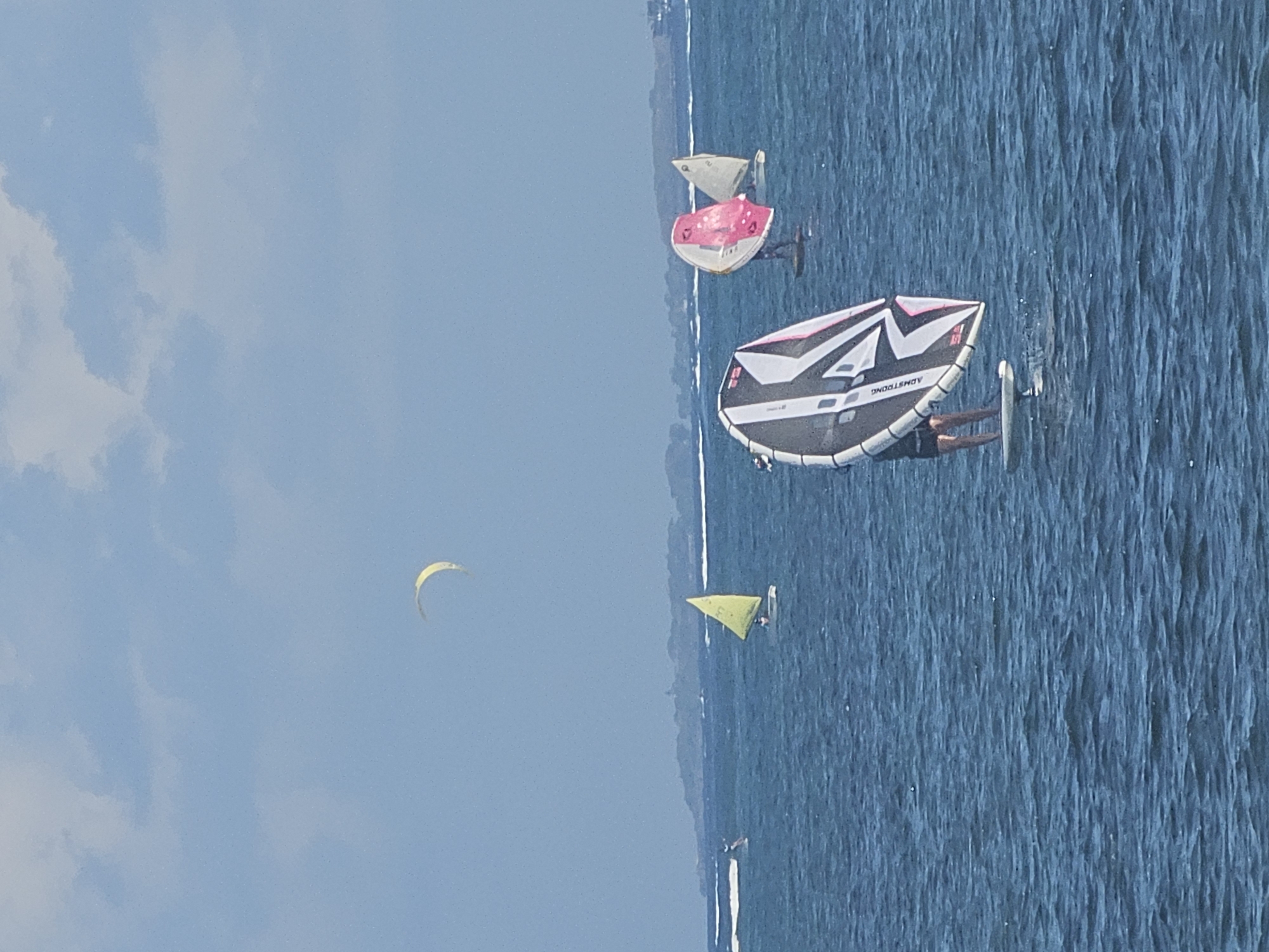 This is an image of people engaged in watersports on a body of water under a partly cloudy sky. In the foreground, a person is on a foilboard, holding a large, black and white sail with pink accents. Further in the distance, several other individuals are visible on smaller sailboats and possibly other wind-powered watercraft. One sailboat has a yellow sail, and another has a pink and white sail. In the far background, a landmass with a hilly coastline is visible. A yellow crescent-shaped object, possibly a kite or part of another sail, is high in the sky. The water appears choppy with small waves. The overall scene depicts a recreational activity on the water with multiple participants. The location is not Denpasar, Indonesia, as the image depicts a coastal scene with a temperate climate and European-style sailboats.