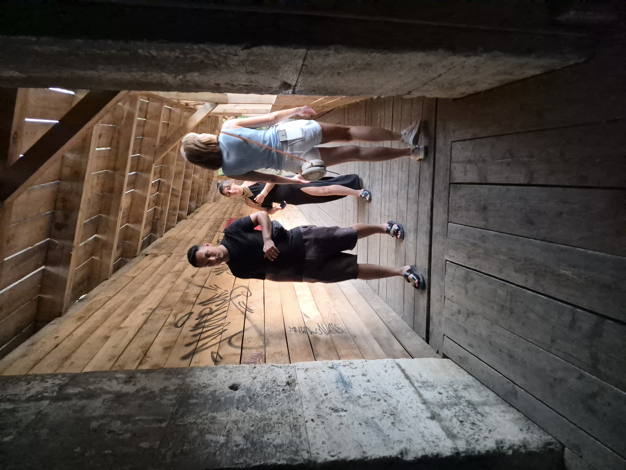 The image depicts a group of three individuals walking down a wooden walkway within a structure constructed of rough-hewn wooden planks, likely part of the city walls in Kotor, Montenegro. The walkway is lined on the left by a rough, concrete wall, possibly a fortification, with graffiti visible on the wooden planks opposite. A man in a black t-shirt and shorts stands facing the camera, holding a small black bag. Behind him, a woman in a long black dress with a slit walks away from the camera. A third individual, a woman wearing a light blue tank top and white shorts, walks ahead of the woman in the black dress, also facing away from the camera, carrying a circular, woven bag. The overhead structure is made of exposed wooden beams and planks, with gaps allowing light to filter through. The floor of the walkway is also made of weathered wooden planks. The scene appears to be an outdoor, historical setting, consistent with the ancient fortifications of Kotor.