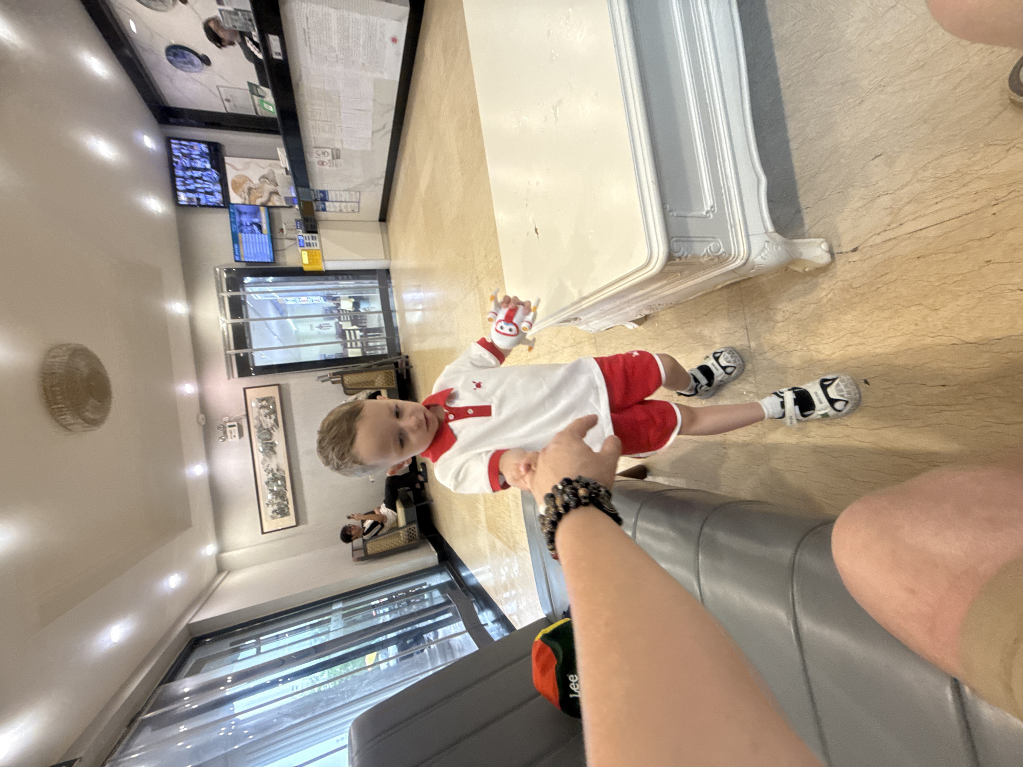 A young boy wearing a white polo shirt with red trim and red shorts is reaching out his hand to an adult in what appears to be a hotel lobby. The boy is holding a small toy in his other hand. In the background, there is a reception desk with computers and staff, as well as seating areas and large windows. The flooring is a light-colored tile. The location is Qinhuangdao, China, as this is a hotel and the image was taken in Qinhuangdao, China.
