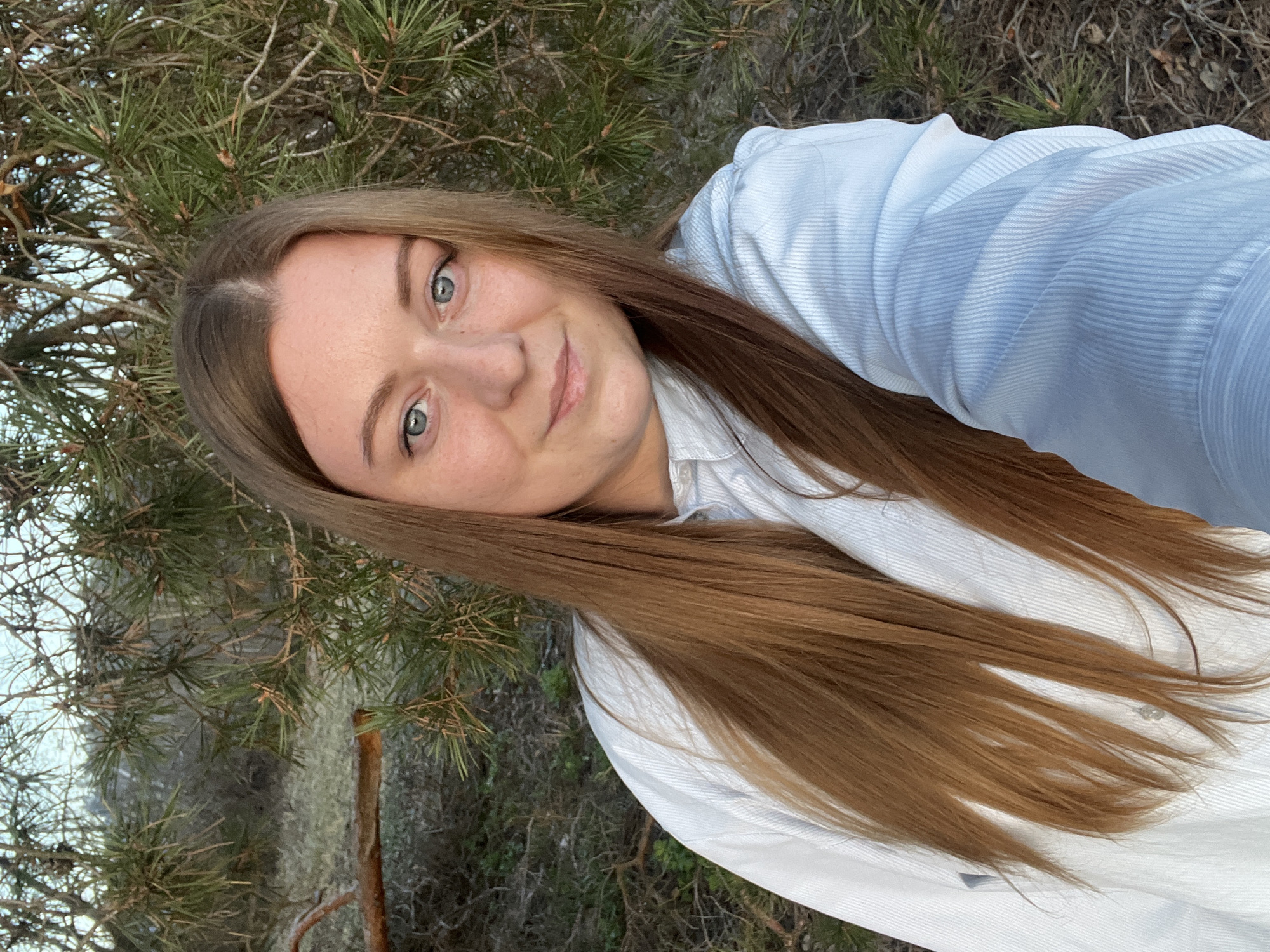 A young woman with long brown hair and blue eyes is pictured in a selfie taken outdoors. She is wearing a light blue, button-down shirt. The background features pine trees and other foliage, suggesting a natural setting. The specific location in Раздолье, Russia, cannot be determined from the image alone.