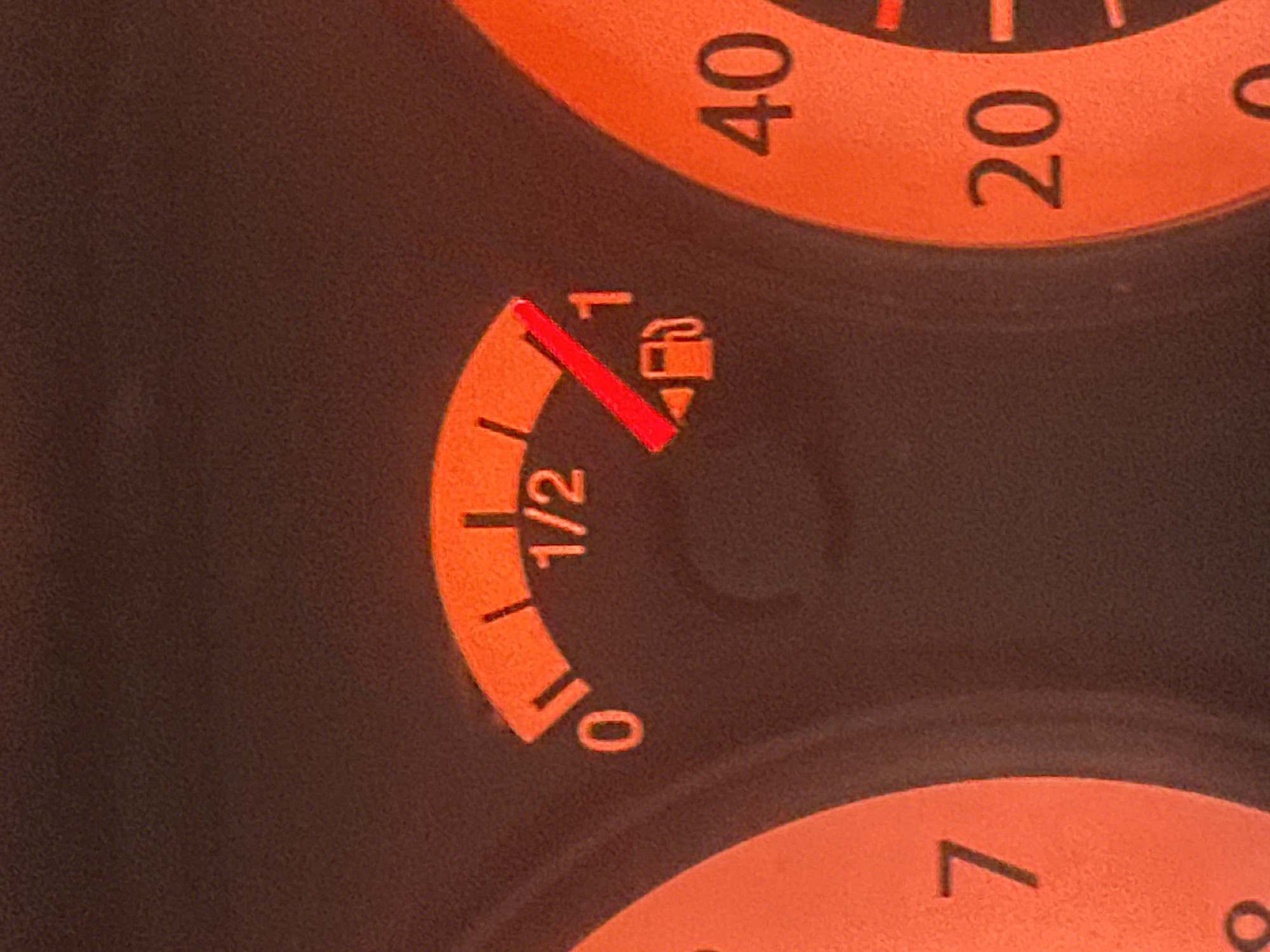 The image shows a close-up of a car's dashboard, specifically the fuel gauge and part of the speedometer. The fuel gauge is illuminated in orange and shows the needle pointing approximately three-quarters of the way between the '1/2' and '1' marks, indicating the fuel tank is more than half full. The markings on the fuel gauge range from '0' to '1'. To the left of the fuel gauge, a portion of another dial is visible with the number '7' at the top. To the right, a section of the speedometer is visible, with the numbers '40' and '20' clearly displayed. The background is a dark, textured surface, likely the dashboard material. The lighting suggests it is dark outside or the car's interior lights are on. There are no people visible in the image, and no information about the location in Tomilova, Russia is present in the visual data.