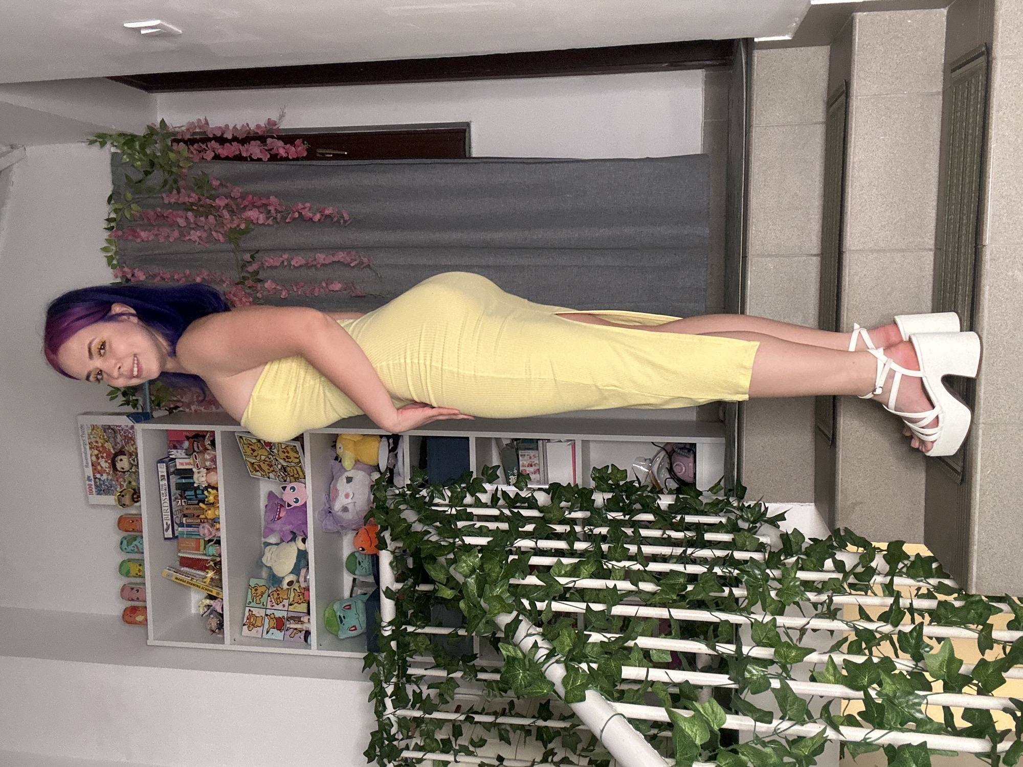 A full-length, rotated color image shows a woman laying atop a white bookshelf decorated with ivy. The woman, who has purple hair, is wearing a yellow dress and white platform heels. She is laying on her side, facing the camera, with one hand placed on her hip and the other down at her side. The bookshelf contains a variety of items, including plush toys, puzzles, books, and figurines. A row of pink flowers lines the top of the bookshelf. The bookshelf sits in front of a grey backdrop and is located to the right of a staircase. The image may have been taken in City of Zagreb, Croatia, but this cannot be confirmed with certainty.