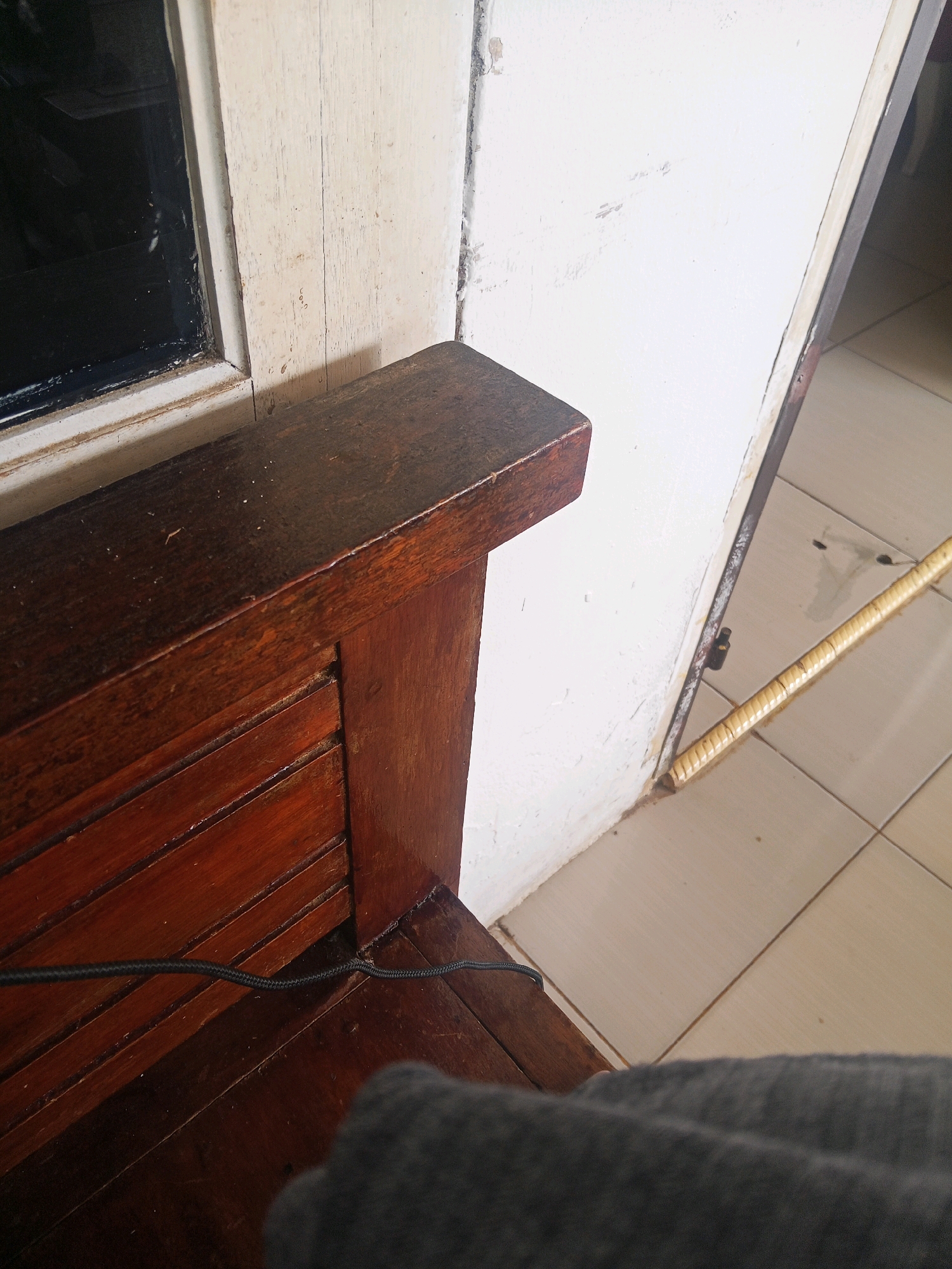 The image shows a close-up view of a wooden bench with a dark finish. The bench has slatted detailing on its backrest. A black cable runs across the slats of the bench. To the right of the bench is a white wall that appears to be partially damaged with scuff marks. A portion of a door frame, painted white and showing signs of wear, is visible. The floor is tiled with light-colored square tiles. A long, thin object with a woven texture, possibly a broom handle, lies on the floor near the wall and door frame. The scene appears to be indoors, in a room with tiled flooring, and the overall impression is one of a functional but somewhat worn living space. The location in Lubuklinggau, Indonesia, suggests a potentially tropical or subtropical climate, which might influence the materials and design of furniture and interior spaces.
