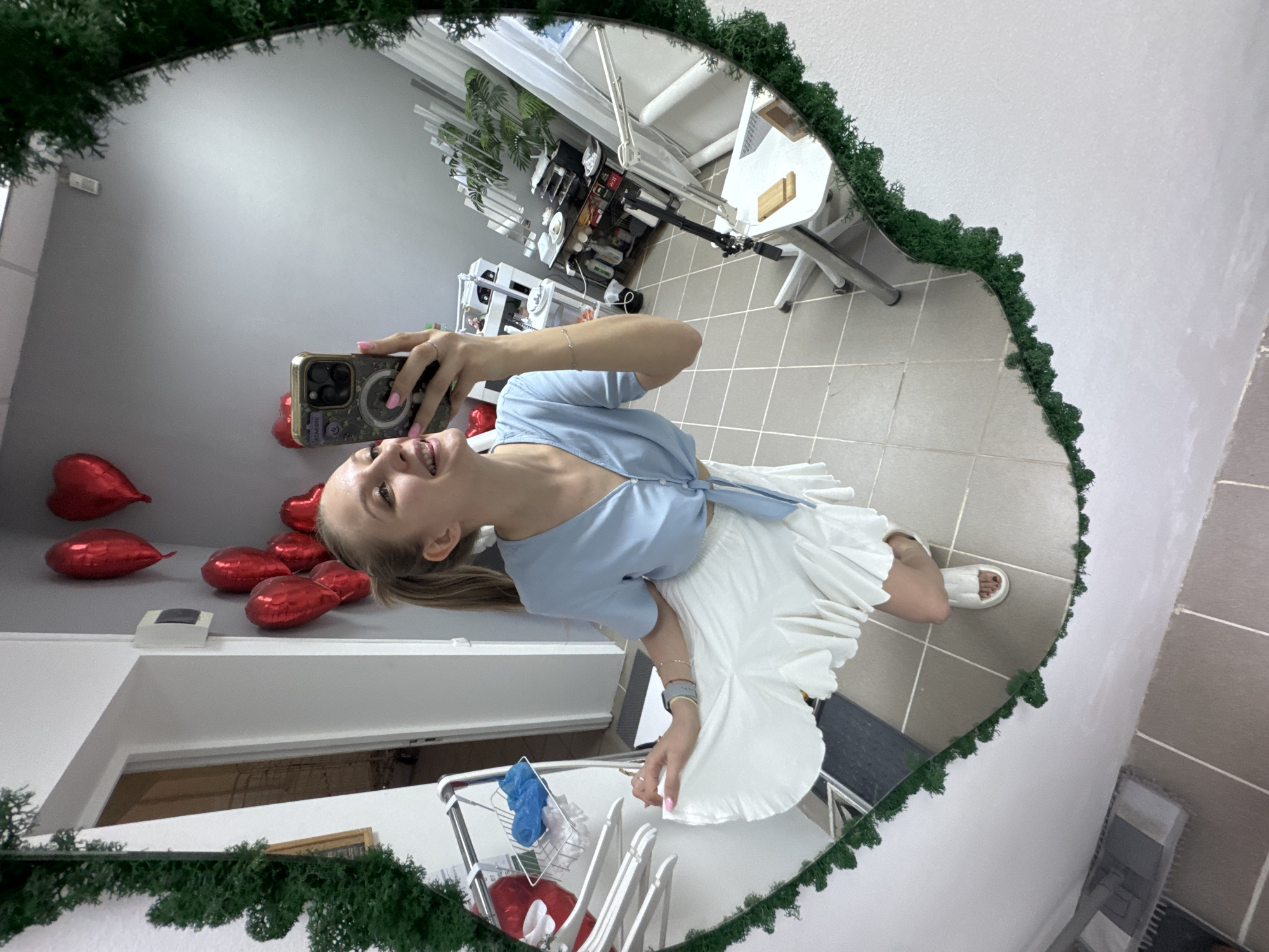 A woman is visible in a mirror selfie, wearing a light blue, tied crop top and a white, pleated skirt, along with white sandals. She has blonde hair pulled back in a ponytail. She is holding a smartphone with a decorative case to take the picture.
In the mirror's reflection, a room is seen decorated with several red heart-shaped balloons attached to the wall. A white table and chairs are present, as well as what appears to be medical or cosmetic equipment in the background, including a medical lamp. There is also a laundry rack with laundry items. The mirror has a decorative green moss frame.
The location is suggested to be in Izhevsk, Russia.