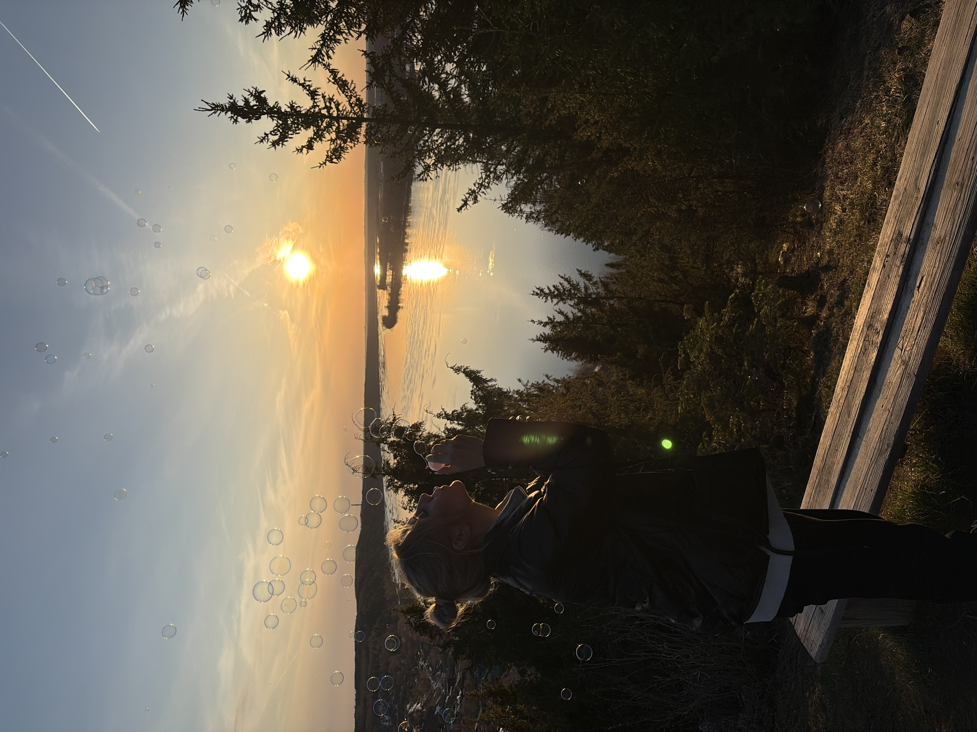 A woman is blowing bubbles while sitting on a wooden bench at dusk, with a body of water and the setting sun in the background. The scene is at least partially set in Ольховка, Russia. The sky transitions from blue to orange near the horizon. A single contrail from an aircraft is visible in the upper-left. The trees along the shore are silhouetted against the light. The woman is wearing dark clothing with a white undershirt, and her hair is blonde. Bubbles are dispersed in front of her and reflecting the light.