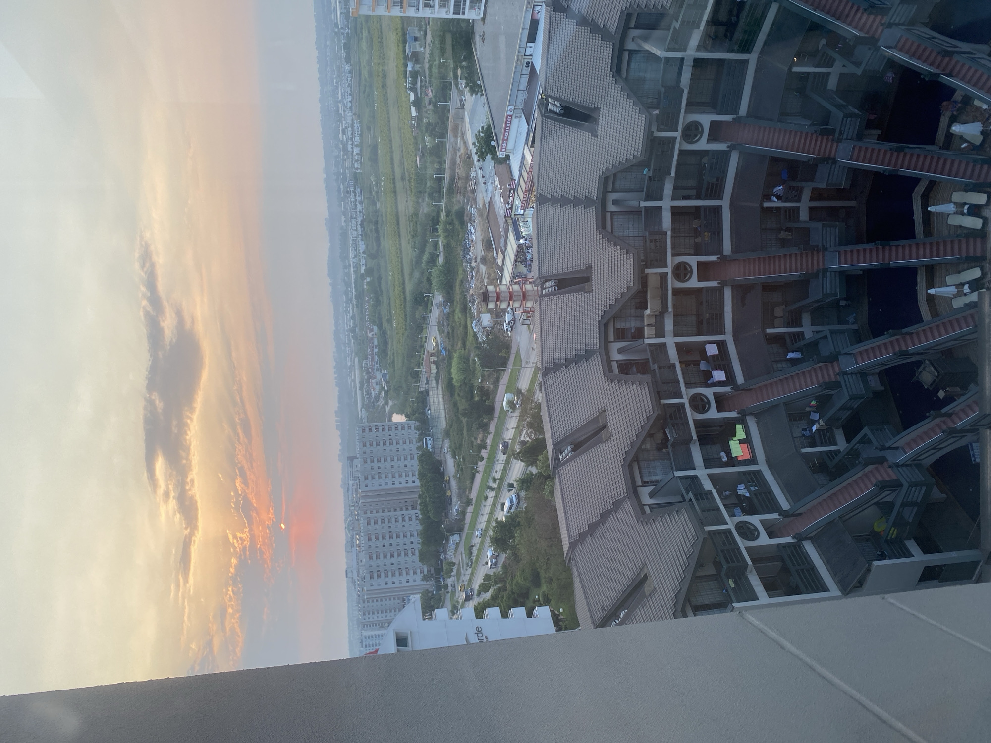 The image is a high-angle view of a resort or hotel complex in Aksu, Turkey, at sunset. The sky is filled with soft hues of orange and yellow, with clouds tinged pink by the setting sun. In the foreground, a distinctive multi-tiered building with brown, angled roofs and numerous balconies is visible. Stairs and walkways connect different levels, and some balconies have clothes hanging to dry. Below, swimming pools and lounge chairs can be seen. Beyond the complex, a street with traffic, including buses and cars, runs alongside a line of apartment buildings and more distant structures. Further into the background, a landscape of green fields and scattered buildings stretches towards a hazy horizon. The overall impression is one of a densely populated urban or resort area under a warm, fading light.
