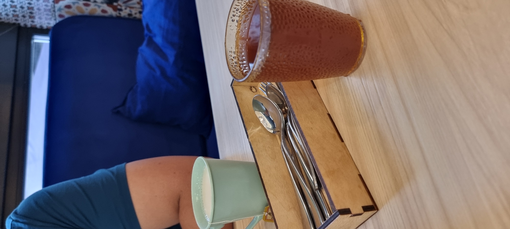 A close-up, eye-level shot shows a light-colored wooden table with various objects on it. In the foreground, a textured glass contains a reddish-brown liquid, likely tea or coffee. To the left, a light green ceramic mug with a tea bag string hanging out holds a milky white beverage. Adjacent to the mug, a wooden organizer tray holds a set of polished metal cutlery, including spoons and forks. In the background, out of focus, is a vibrant blue sofa with a dark blue cushion, and a portion of a person's arm wearing a teal t-shirt. The scene suggests a casual indoor setting, potentially a cafe or restaurant. No specific landmarks or context are visible to pinpoint a location within Almaty, Kazakhstan.