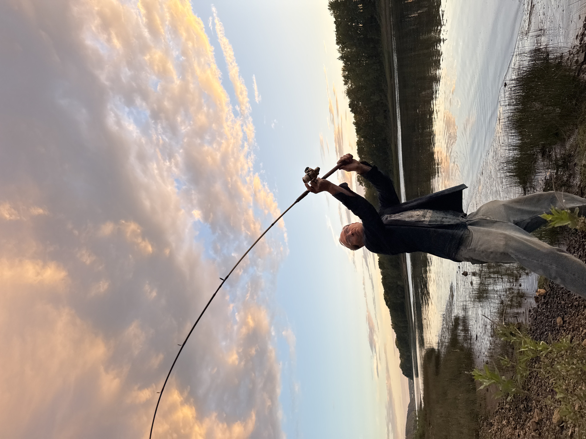 A man is fishing on the bank of a river at dusk. He is standing and casting his line with his fishing rod. The sky is filled with colorful clouds, a mixture of pink, orange, and blue. The river reflects the sky and is surrounded by a treeline on the opposite bank. The foreground shows the muddy bank with some vegetation.