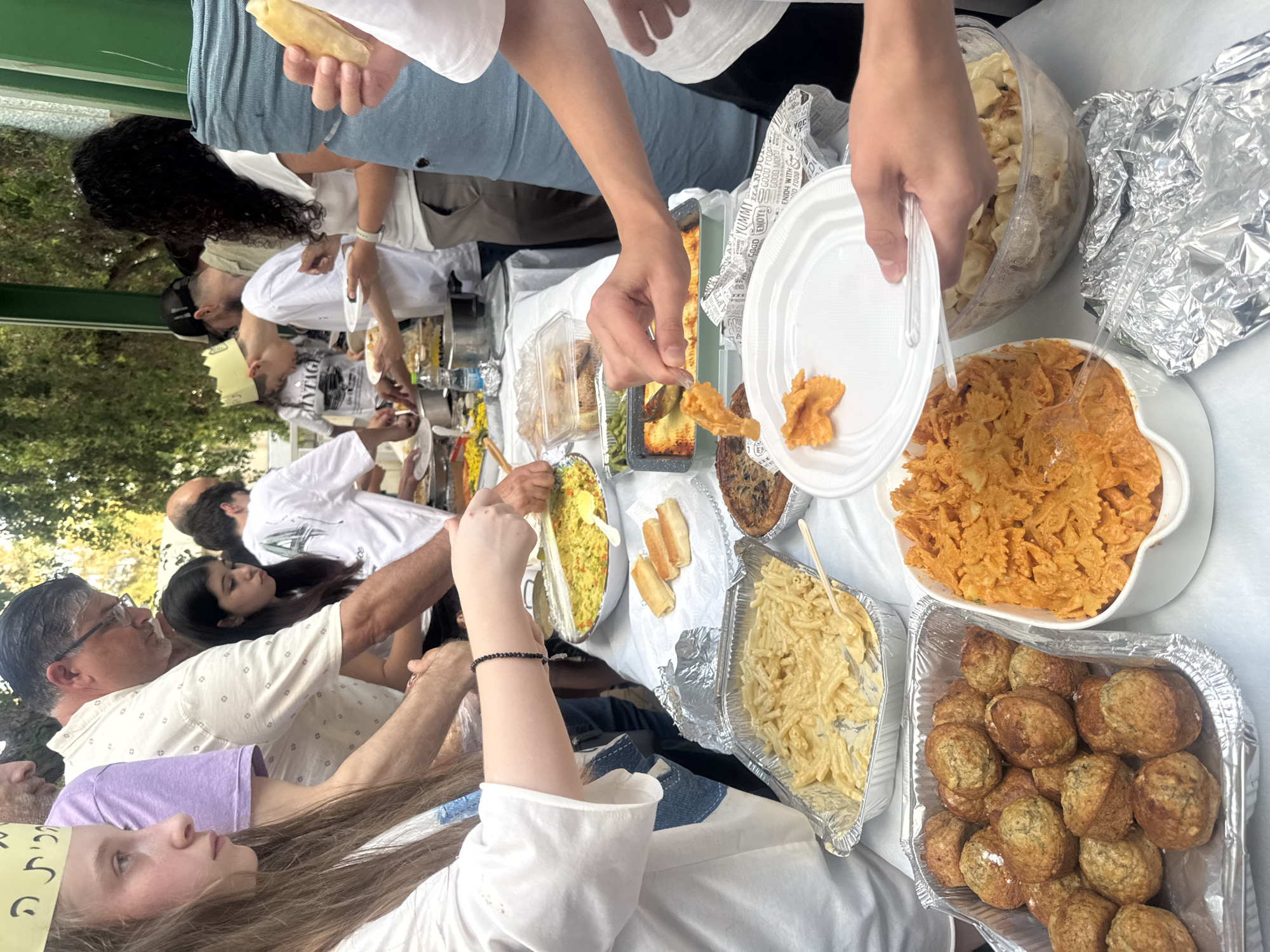 This is a medium shot, overhead, slightly angled view of a group of people gathered around a table laden with food. The scene appears to be an outdoor event, possibly a picnic or a family gathering, with greenery and trees visible in the background, suggesting a park or backyard setting. The lighting indicates it's daytime, likely sunny.
Several individuals are visible, with a focus on their hands interacting with the food. One person is serving themselves a piece of fried food from a platter, while others are reaching for various dishes. The table is filled with diverse food items, including pasta dishes (macaroni and cheese, and a red-sauced pasta), what looks like rice or couscous, baked goods, meatballs or falafel, and what appears to be a dish of dumplings or tortellini. Some of the food is served in disposable aluminum trays and plastic containers, while other items are in larger bowls. There are also disposable plates and cutlery.
The people are dressed casually, with t-shirts and light clothing visible, reinforcing the outdoor, informal atmosphere. The overall mood seems convivial and relaxed, with people enjoying a meal together.
Visible text includes "FOR YUMMY," "GOGO ENOY!," and "GOOD FOOD," printed on paper napkins scattered among the food. There is also Hebrew text on a yellow band worn by one of the children, but it is partially obscured and difficult to read. The location context of Givatayim, Israel, suggests a potentially Israeli or Middle Eastern cultural context for the gathering and food.