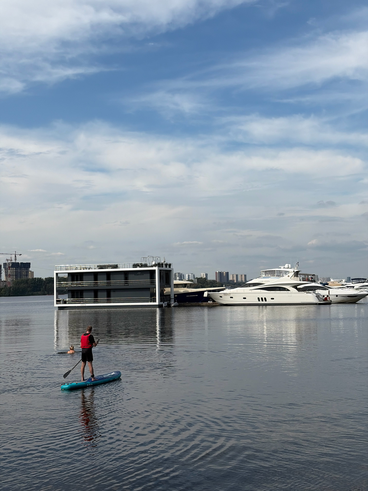 The image depicts a waterfront scene in Khimki, Russia, on a cloudy day. In the foreground, a person wearing a red life vest and black shorts stands on a teal paddleboard, holding a paddle. To the left and further back in the water, another person is swimming. In the mid-ground, a modern, two-story houseboat with a white frame and dark gray exterior is moored. Further to the right, a large white yacht with multiple decks and windows is docked. In the background, the cityscape of Khimki is visible, with several multi-story buildings and construction cranes. The water is calm with ripples, reflecting the sky and the structures on the shore.